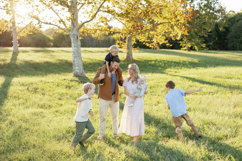 Happy family with children playing in a sunny green meadow surrounded by autumn trees during golden hour.