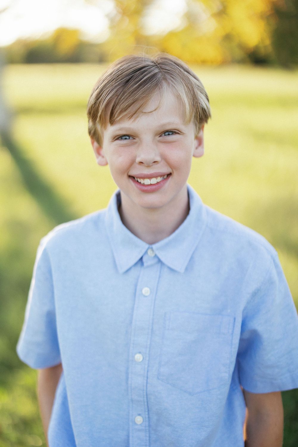 Smiling blond boy in a light blue button-up shirt posing outdoors in a sunny green field.