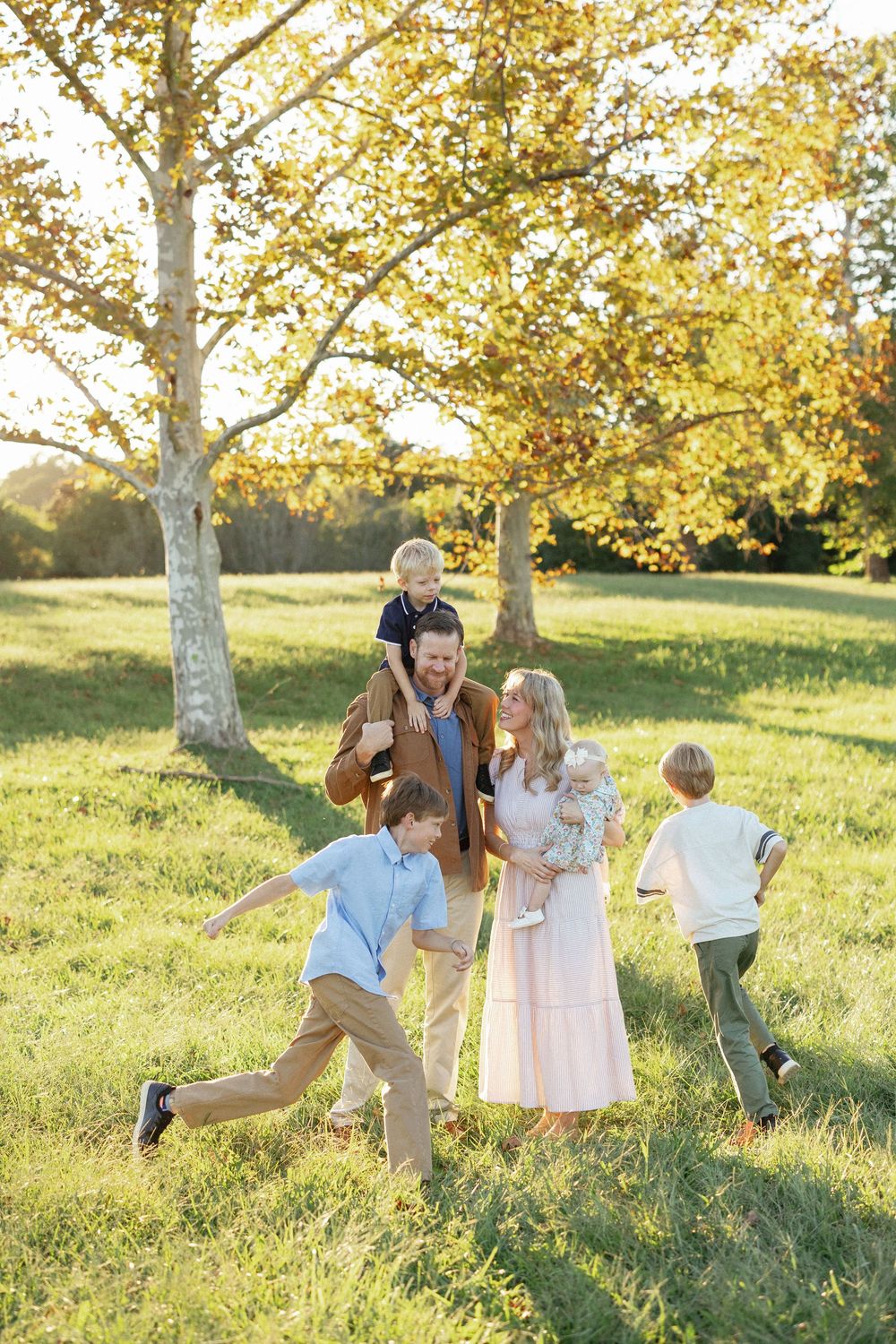 Family of five enjoying a sunny autumn day outdoors on green grass with golden trees in the background.
