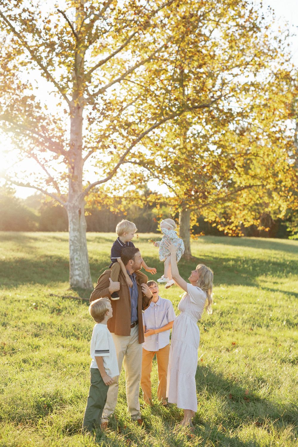 Family of five enjoying a sunny autumn day in the park, with father holding son on shoulders and mother lifting baby.