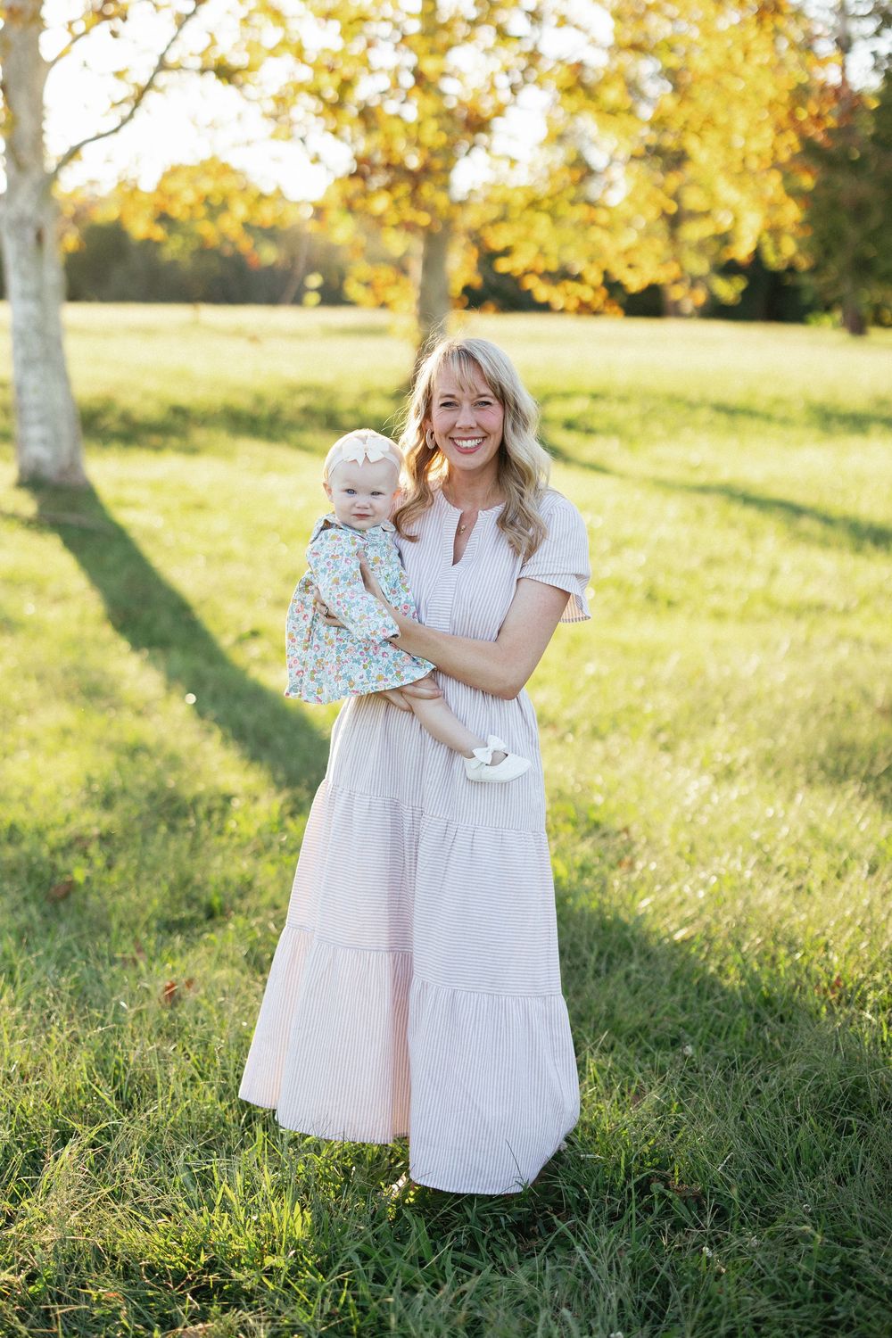 Smiling blonde woman in pink maxi dress holding baby girl in floral outfit, standing in sunny green park.