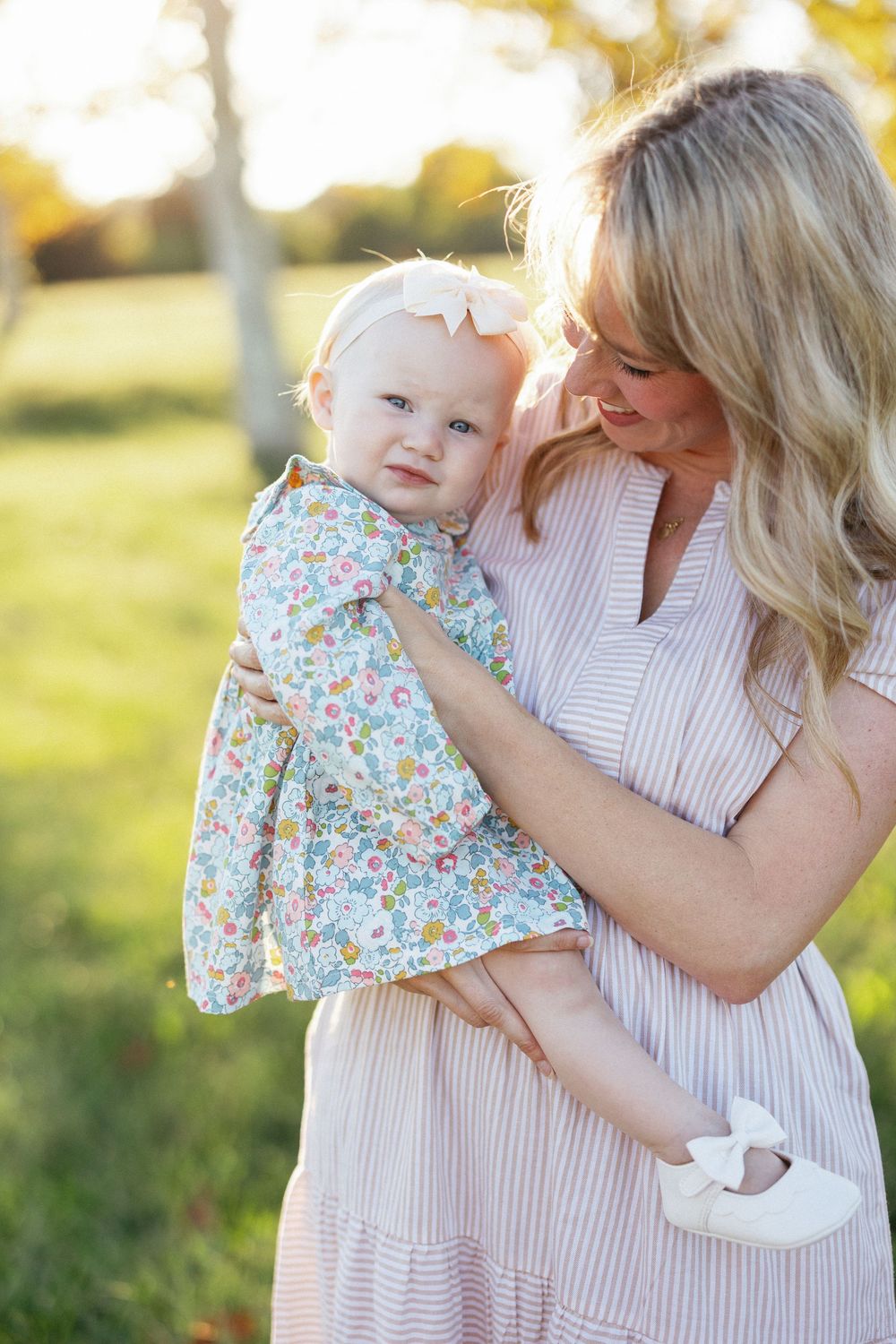 A blonde mother in a striped dress holds her baby girl in a floral outfit with a pink bow outdoors.