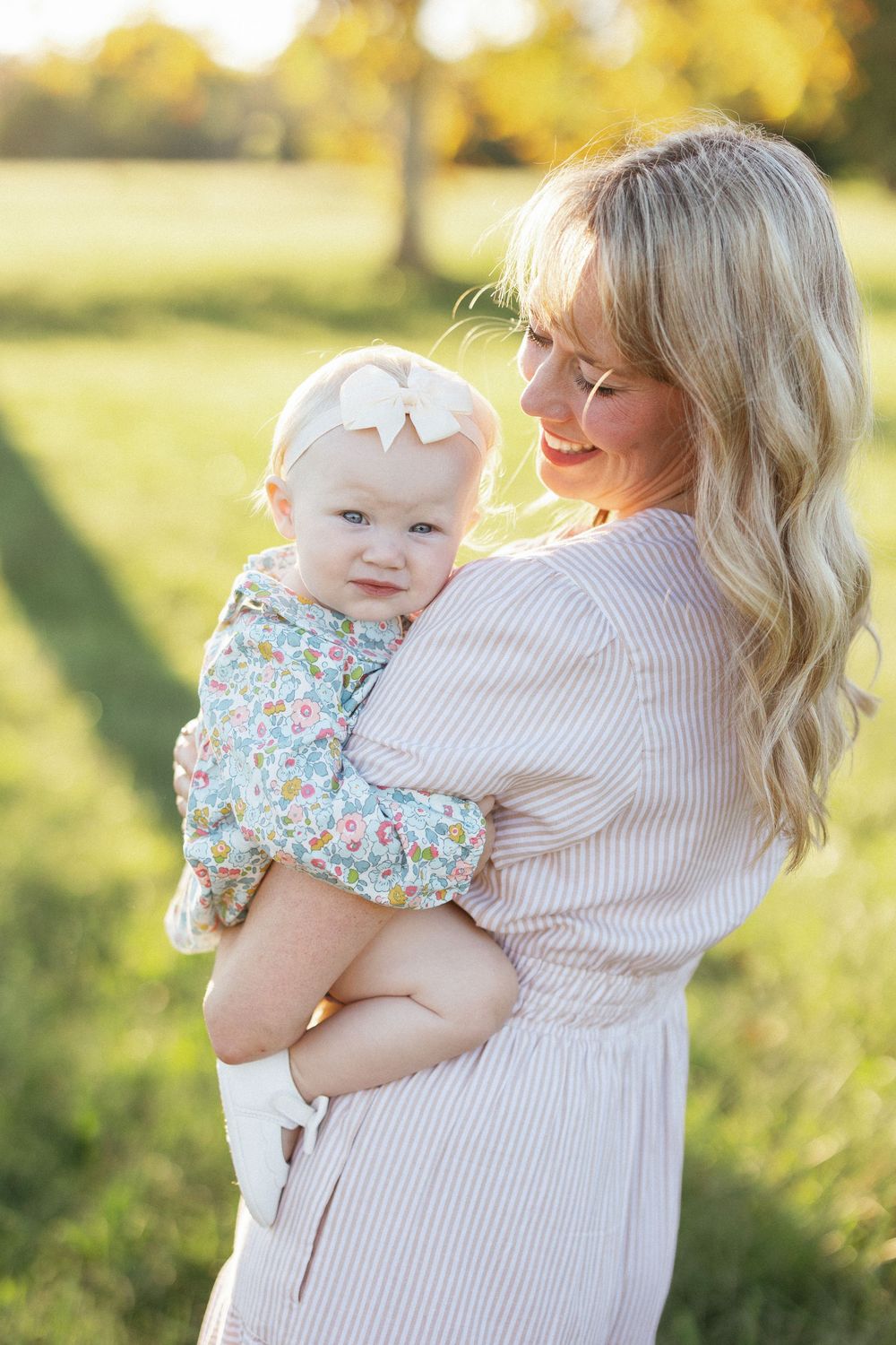 Smiling blonde mother holding her baby girl in a floral outfit with a bow headband outdoors in a sunny green field.