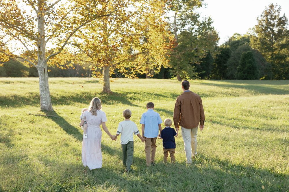 Family of five walking hand-in-hand through a sunny green meadow with golden autumn trees in the background.