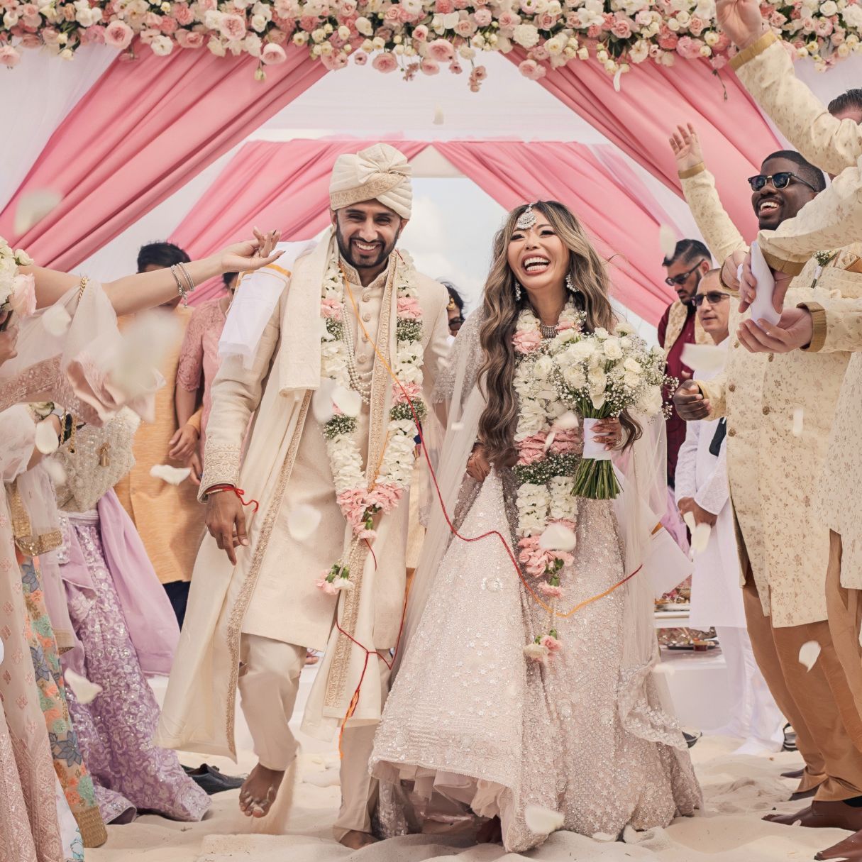 A joyful couple in traditional Indian wedding attire walks under a floral arch, surrounded by celebrating guests.