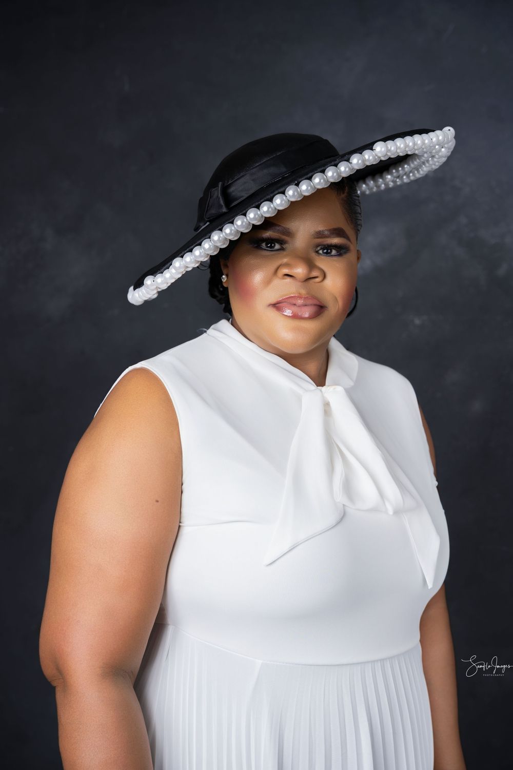 A woman in a white sleeveless dress and elegant black wide-brim hat adorned with pearls poses for a professional portrait.