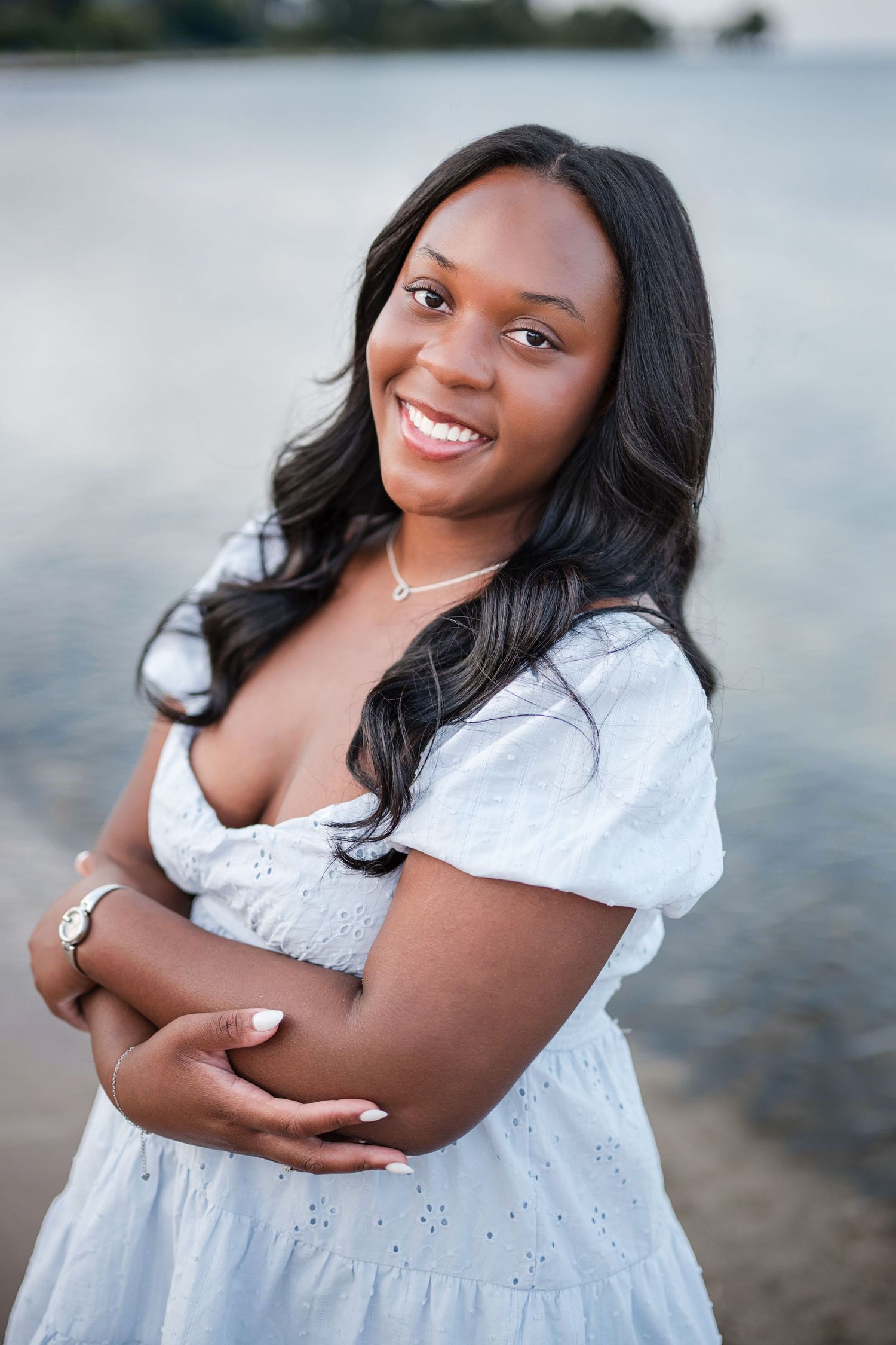 Smiling young Black woman in white dress with arms crossed, posing outdoors near water.