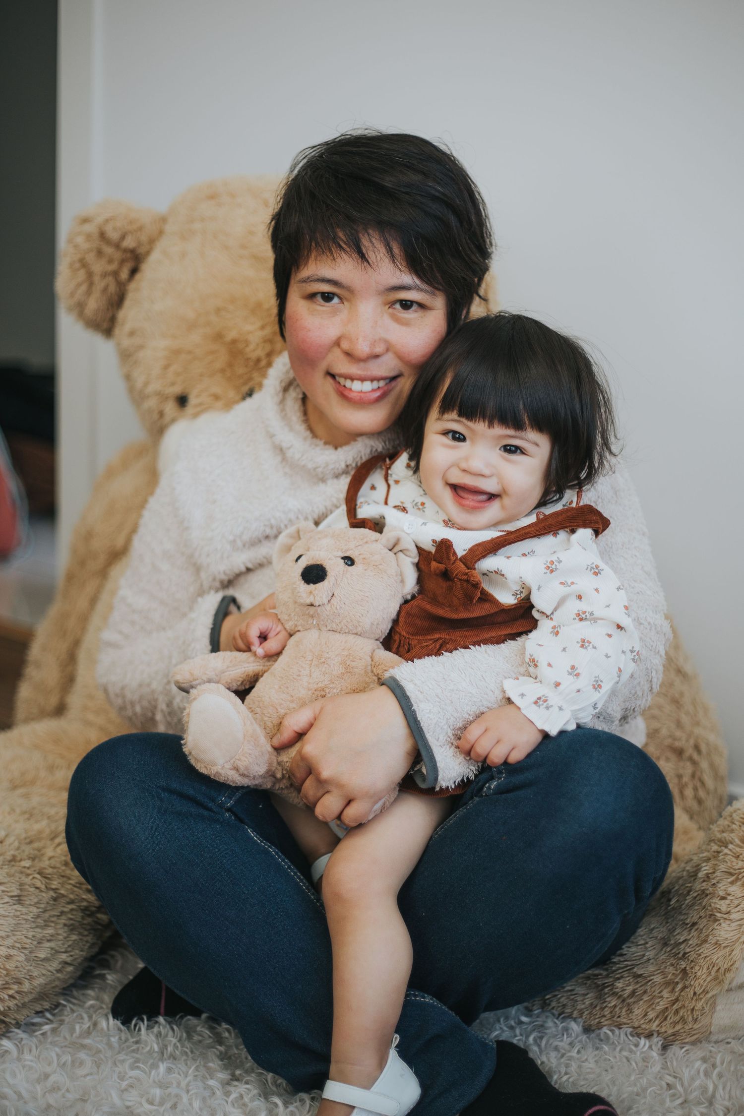 A smiling woman holds a happy toddler and small teddy bear, seated in front of a large plush bear.