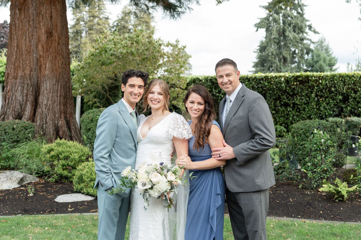 Wedding party of four posing outdoors, bride holding white bouquet, wearing lace gown, flanked by guests in blue and gray suits.