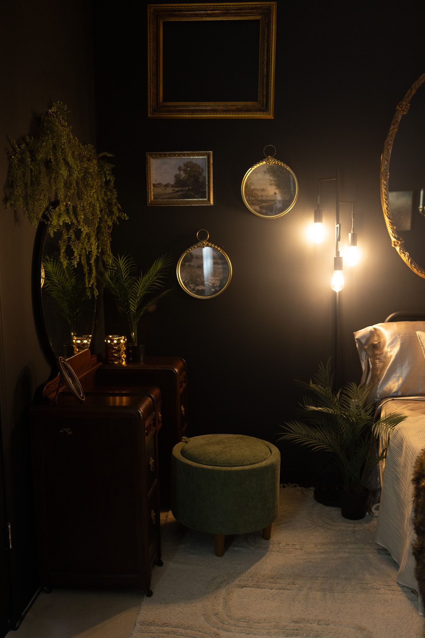 Dark moody bedroom with green velvet ottoman, vintage mirrors, gold frames, lush plants, and warm glowing wall sconce light.