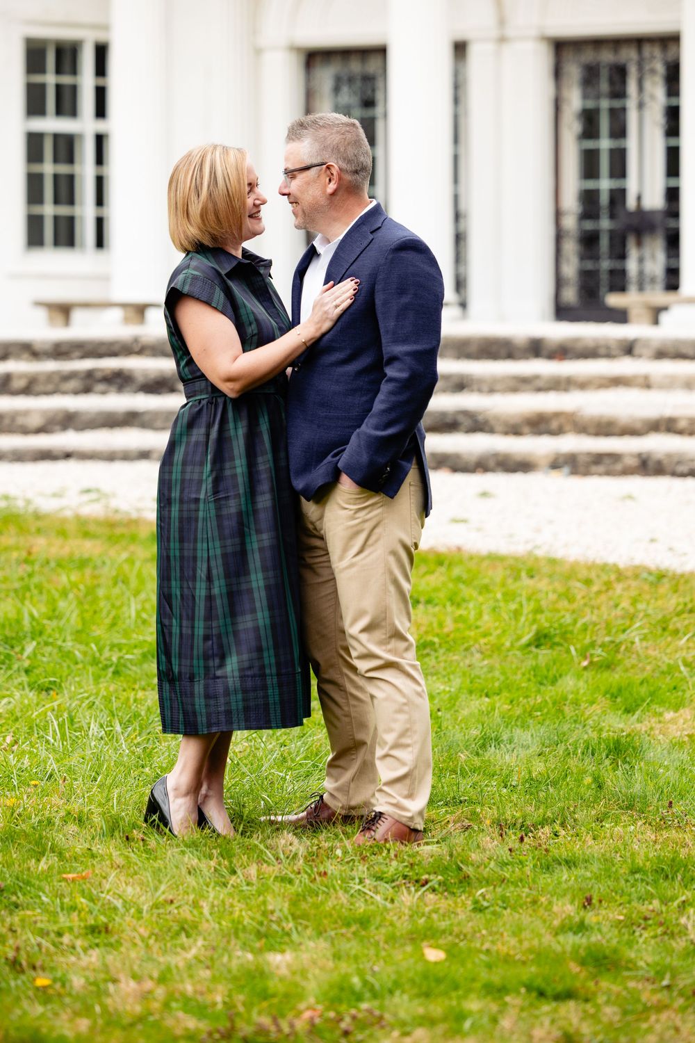 Middle-aged couple sharing a romantic moment on green lawn outside elegant white building, woman in plaid dress, man in navy blazer.