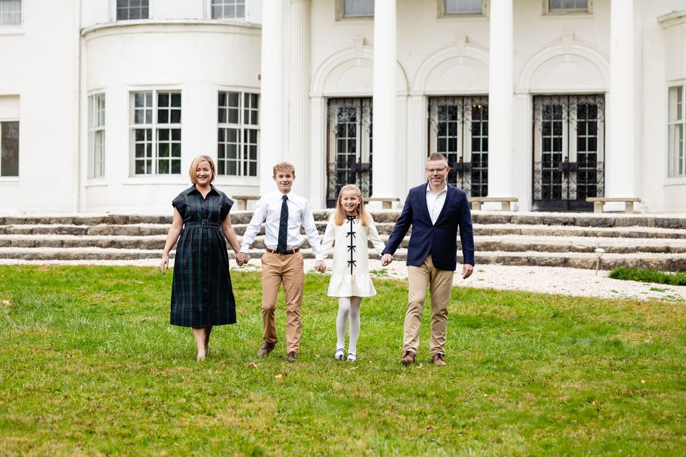 Family of four holding hands walking on green lawn in front of elegant white mansion with columns.