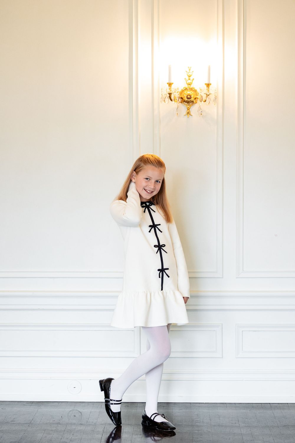 A smiling young girl in a white dress with black bows and white tights poses by an elegant wall sconce.