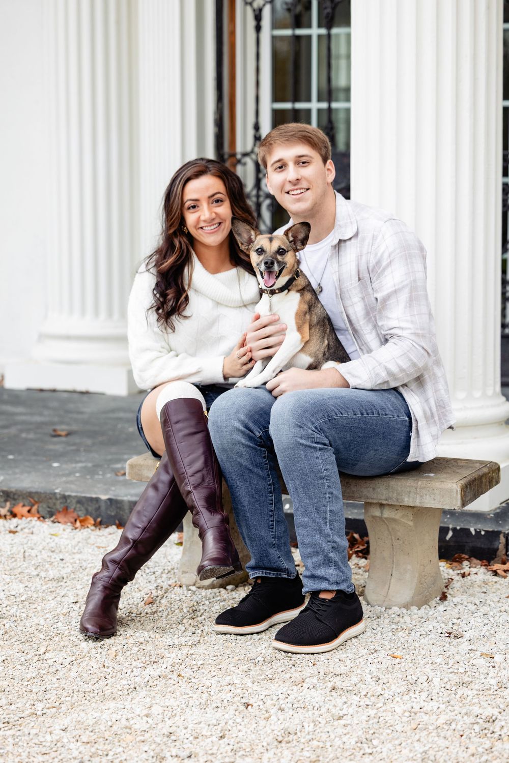 A smiling couple sits on a stone bench holding a small dog in front of a white columned building.