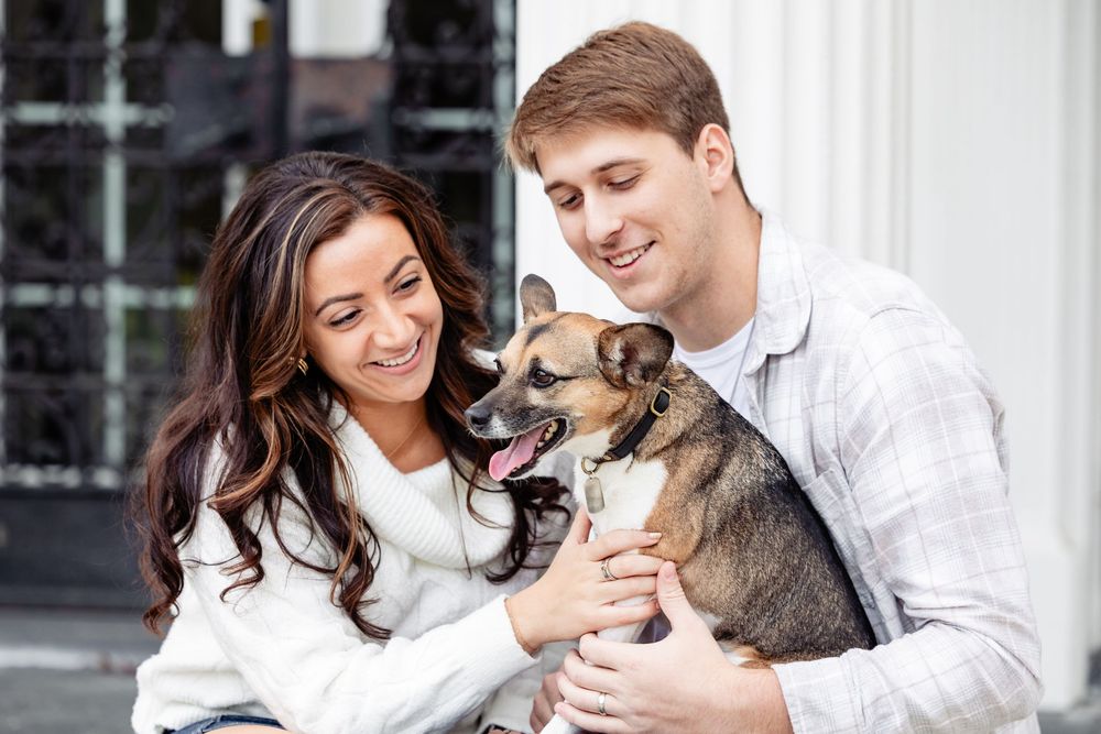 Happy couple smiling and holding an adorable dog outdoors, sharing a joyful moment together.