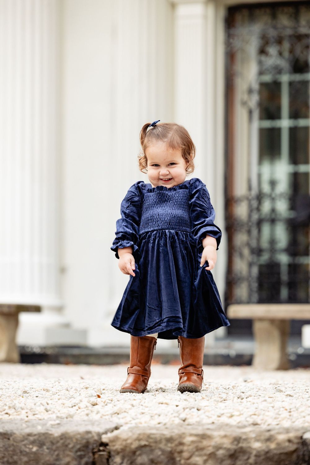 Toddler girl in navy blue velvet dress and brown cowboy boots smiling outdoors near white columns.