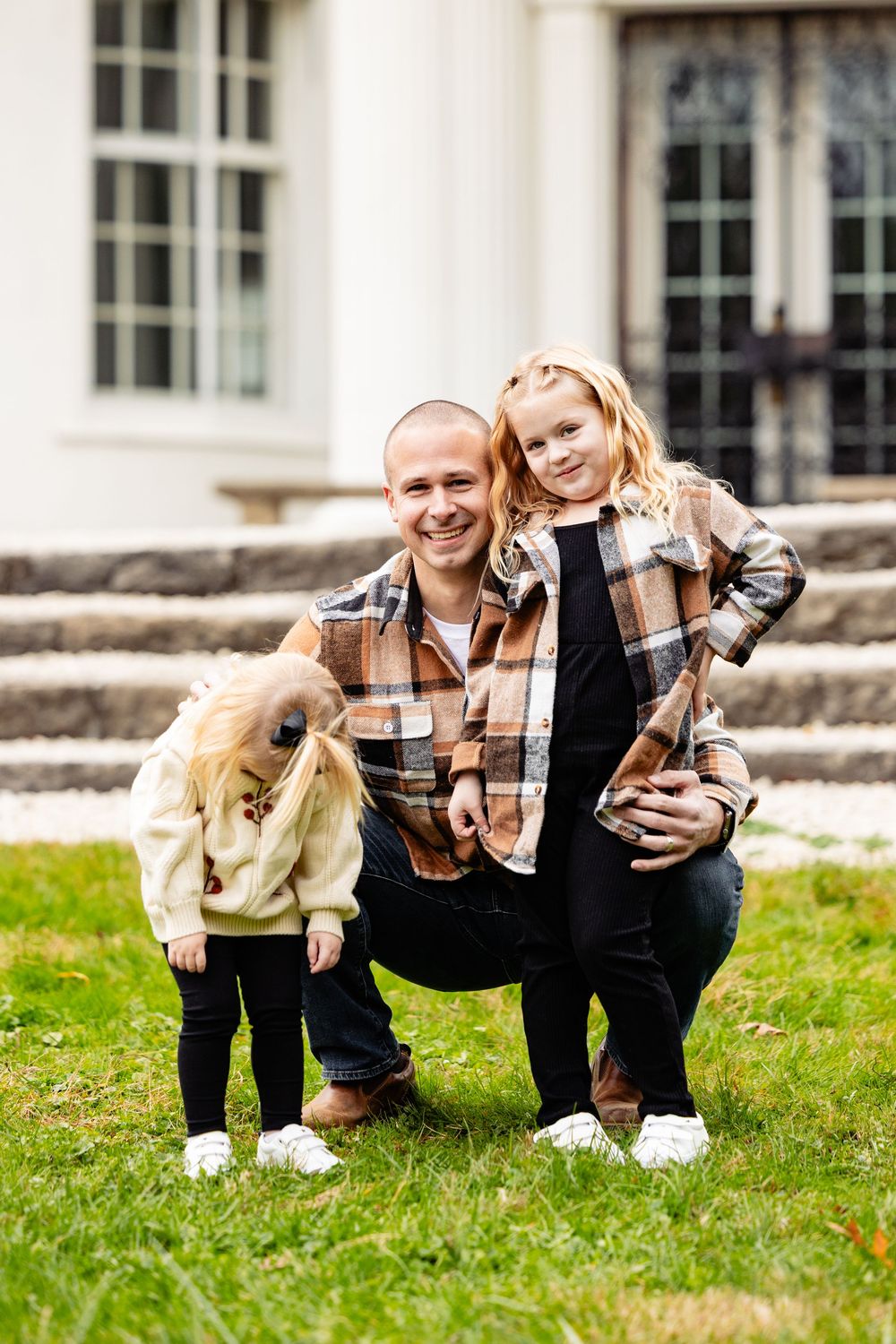 A smiling father crouches on green grass with two young blonde daughters in matching plaid shirts outside a white house.