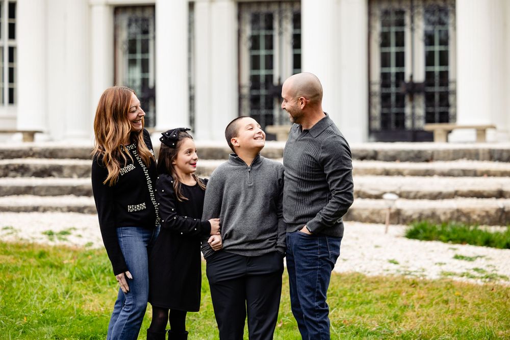 Happy family of four laughing together outdoors in front of a white colonial building on a green lawn.