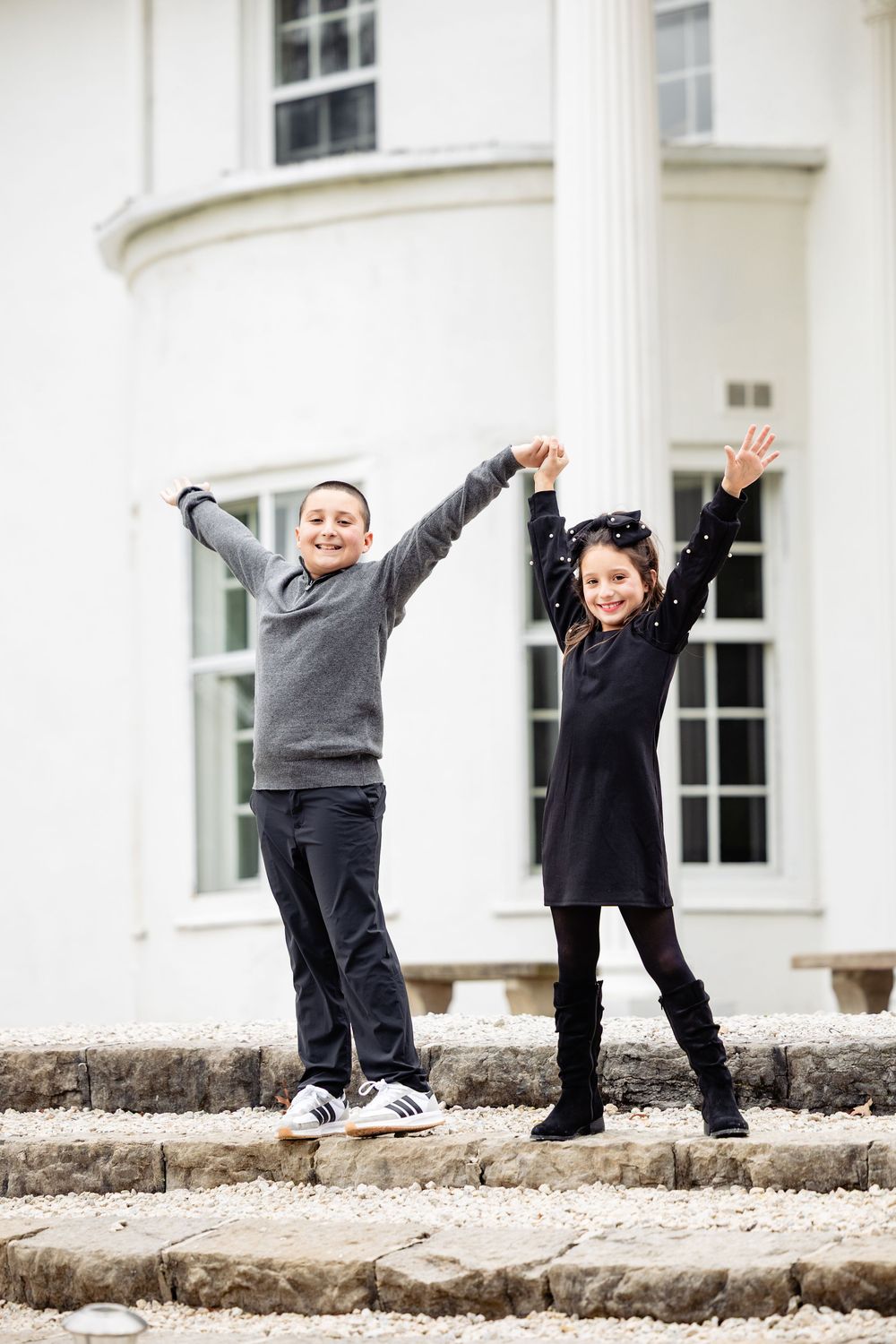 Two children joyfully raising their arms outside a white building, wearing gray and black outfits on stone steps.
