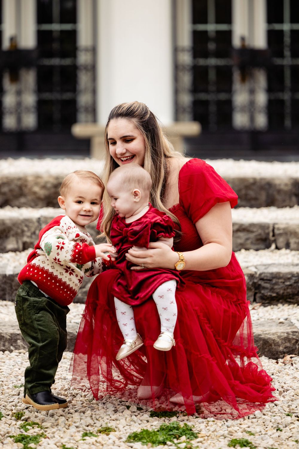 A smiling woman in a red dress holds a baby girl while a toddler boy in a holiday sweater poses outdoors.