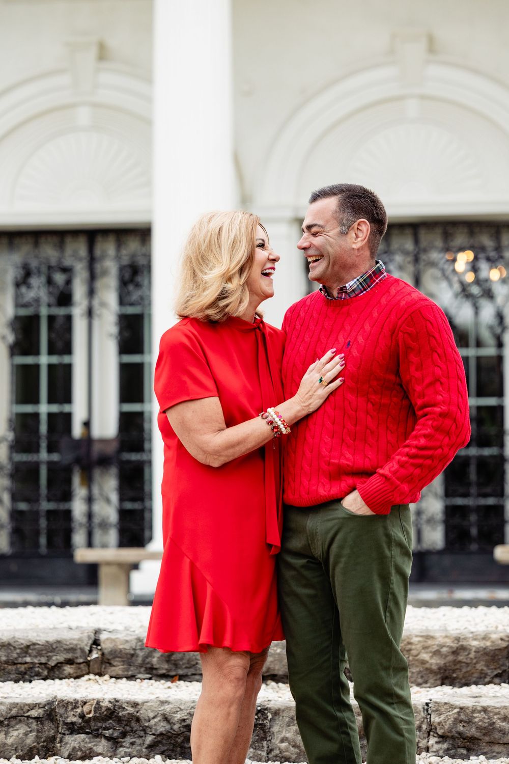 A smiling middle-aged couple in matching red outfits embracing outdoors in front of a white columned building.