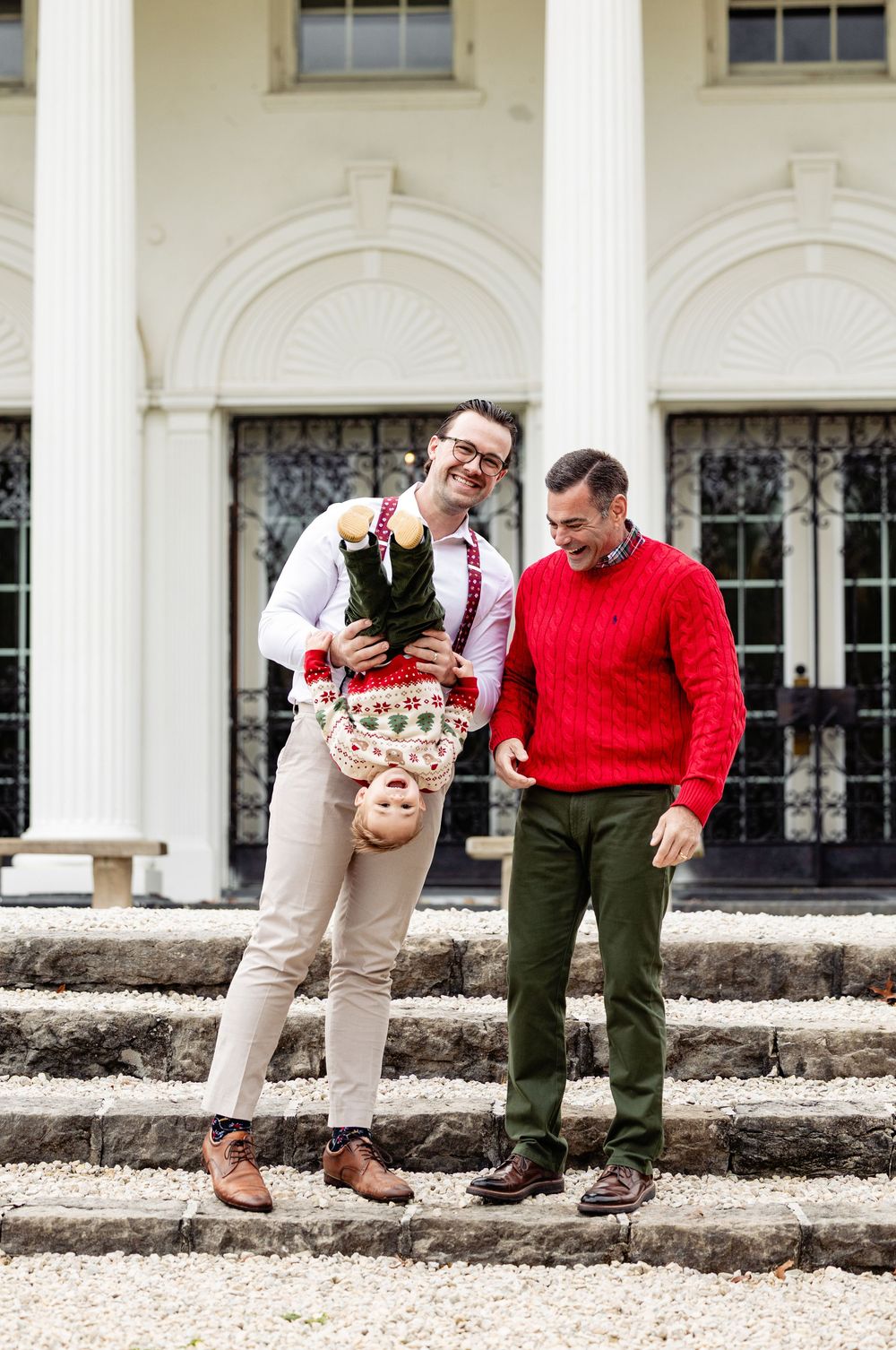 Two men laughing outdoors, one holding a child upside down, wearing festive holiday sweaters on building steps.