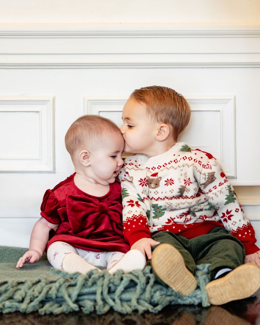 Toddler boy in Christmas sweater kissing baby girl in red velvet dress while sitting on green blanket.