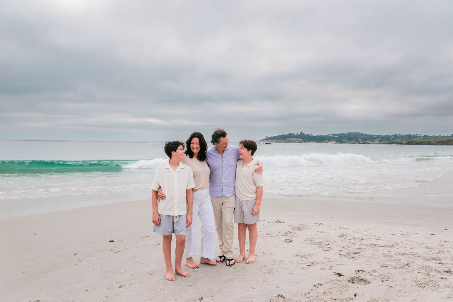 Family of four smiling together on a sandy beach with turquoise waves and an overcast sky. Carmel-by-the-Sea family photographer