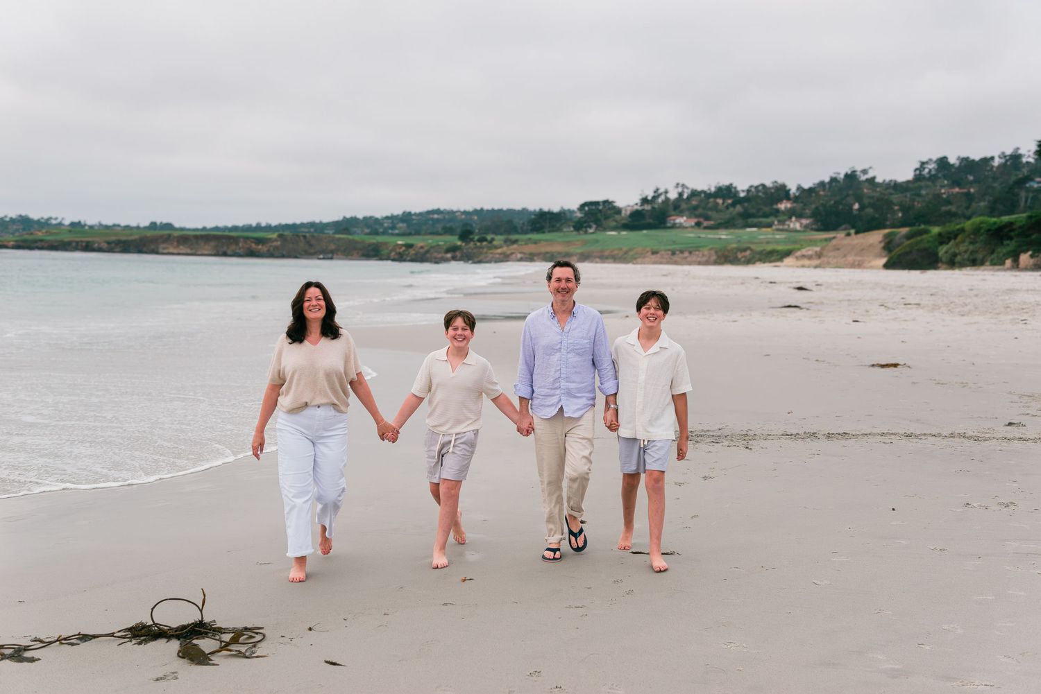 Family holding hands walking on a sandy beach with calm ocean waves and green trees in the background. Carmel-by-the-Sea family photographer