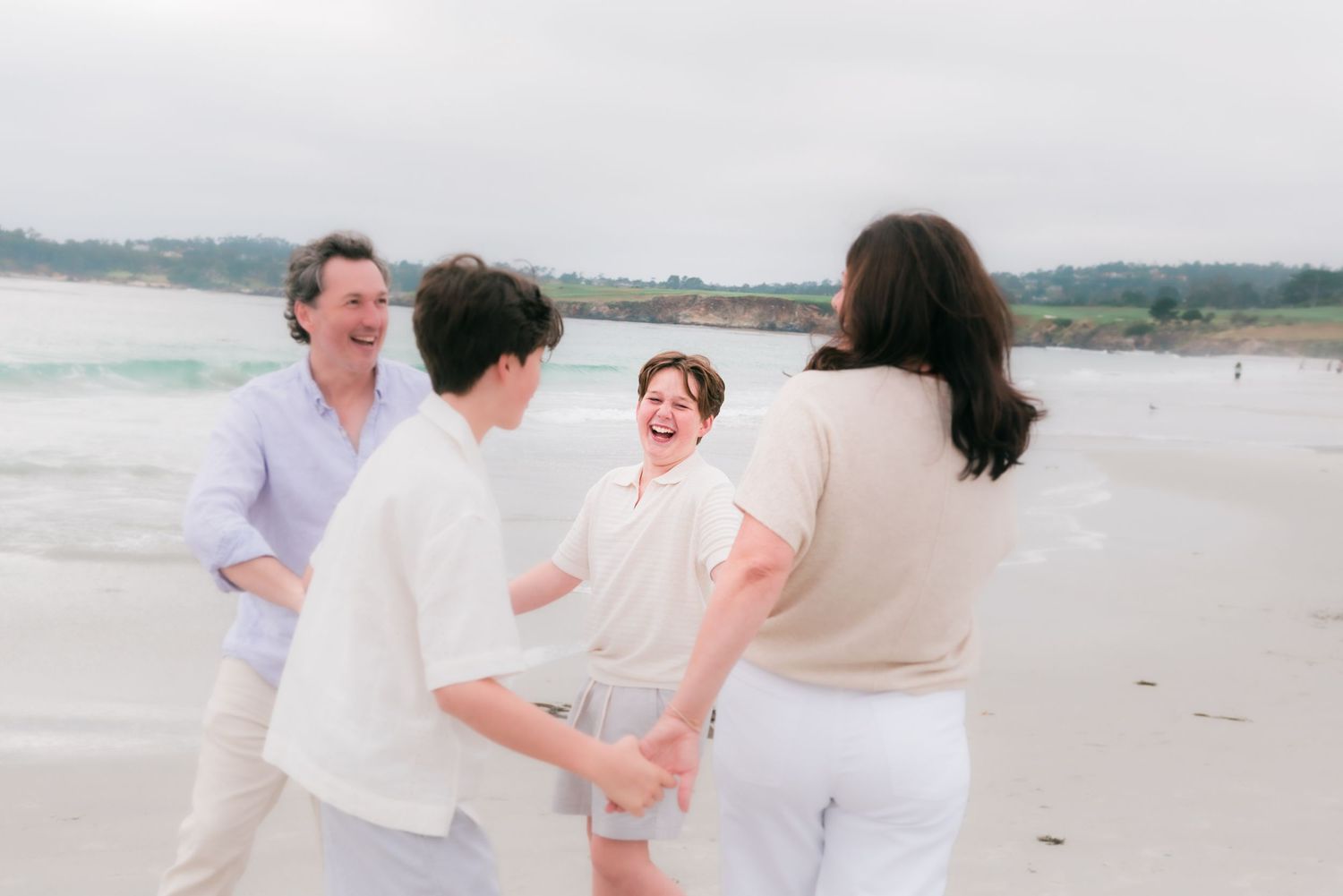Happy family laughing and playing together on a sandy beach with ocean waves in the background. Carmel-by-the-Sea family photographer