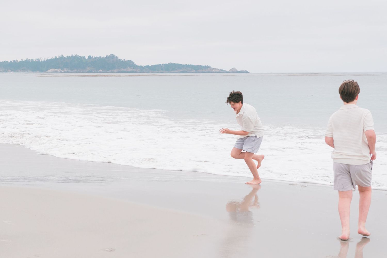 children playing at the ocean shore on a cloudy beach day, with waves and a distant island visible. Carmel-by-the-Sea family photographer