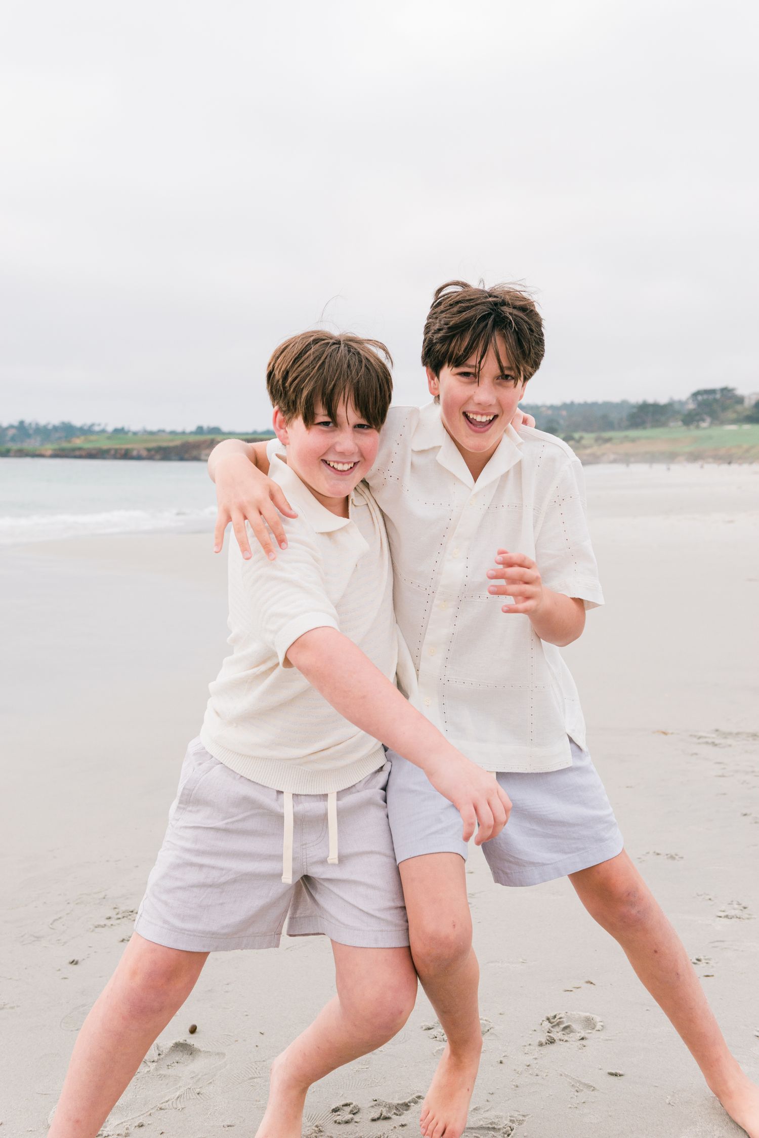 Two happy boys in white shirts and gray shorts laugh and pose together on a sandy beach. Carmel-by-the-Sea family photographer