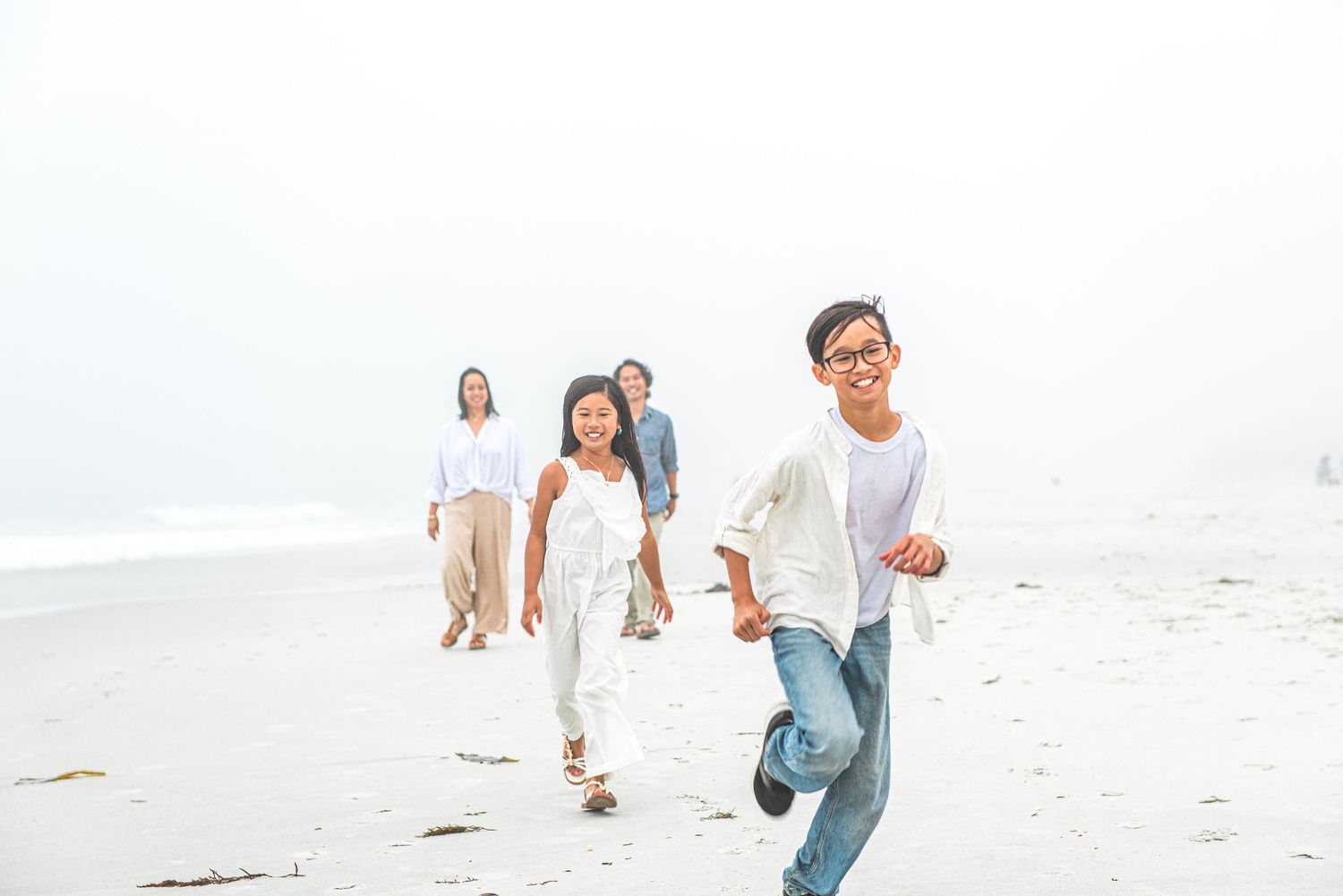 Family walking on a misty beach, with a smiling boy in glasses running ahead of his parents and sister.Carmel-by-the-Sea family photographer