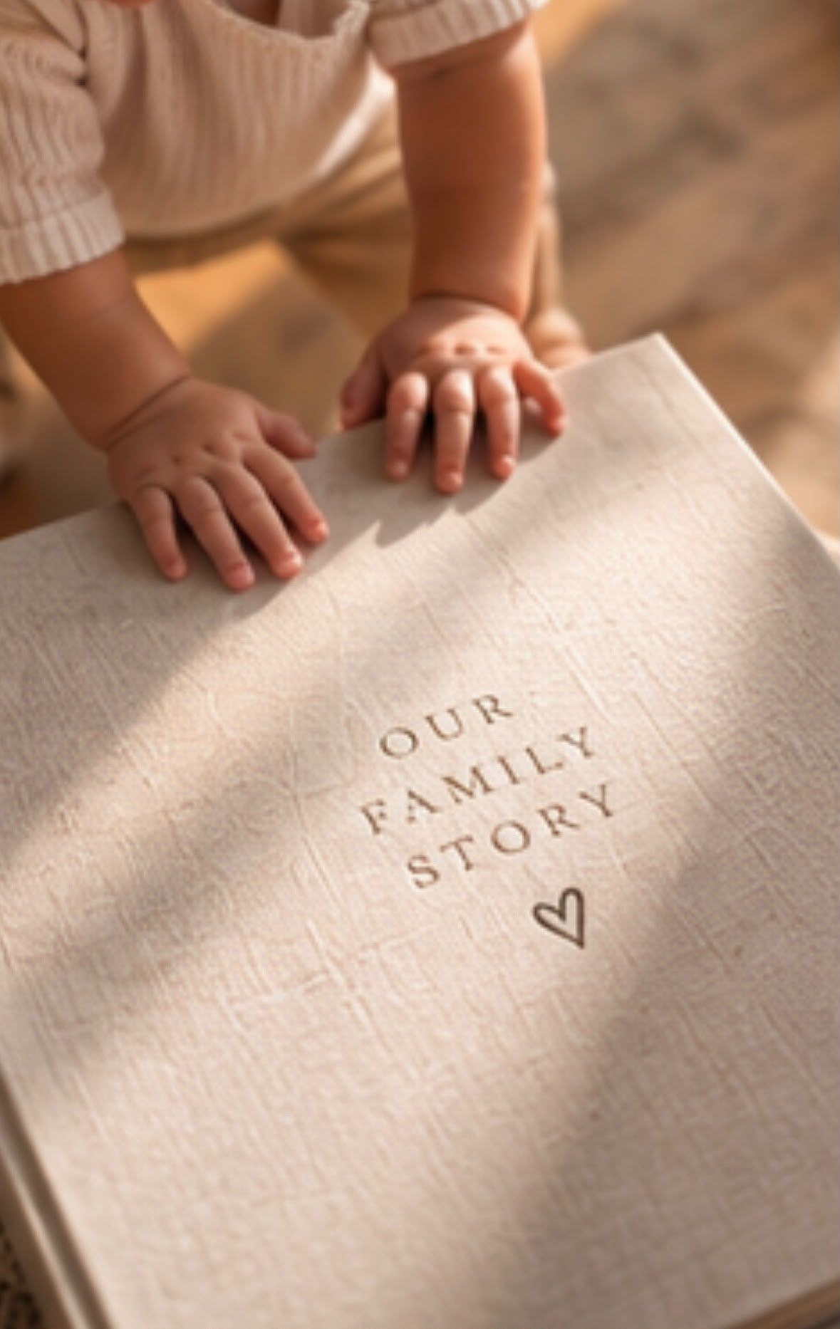 A baby's hands rest on a linen-covered Our Family Story keepsake book with a heart emblem.