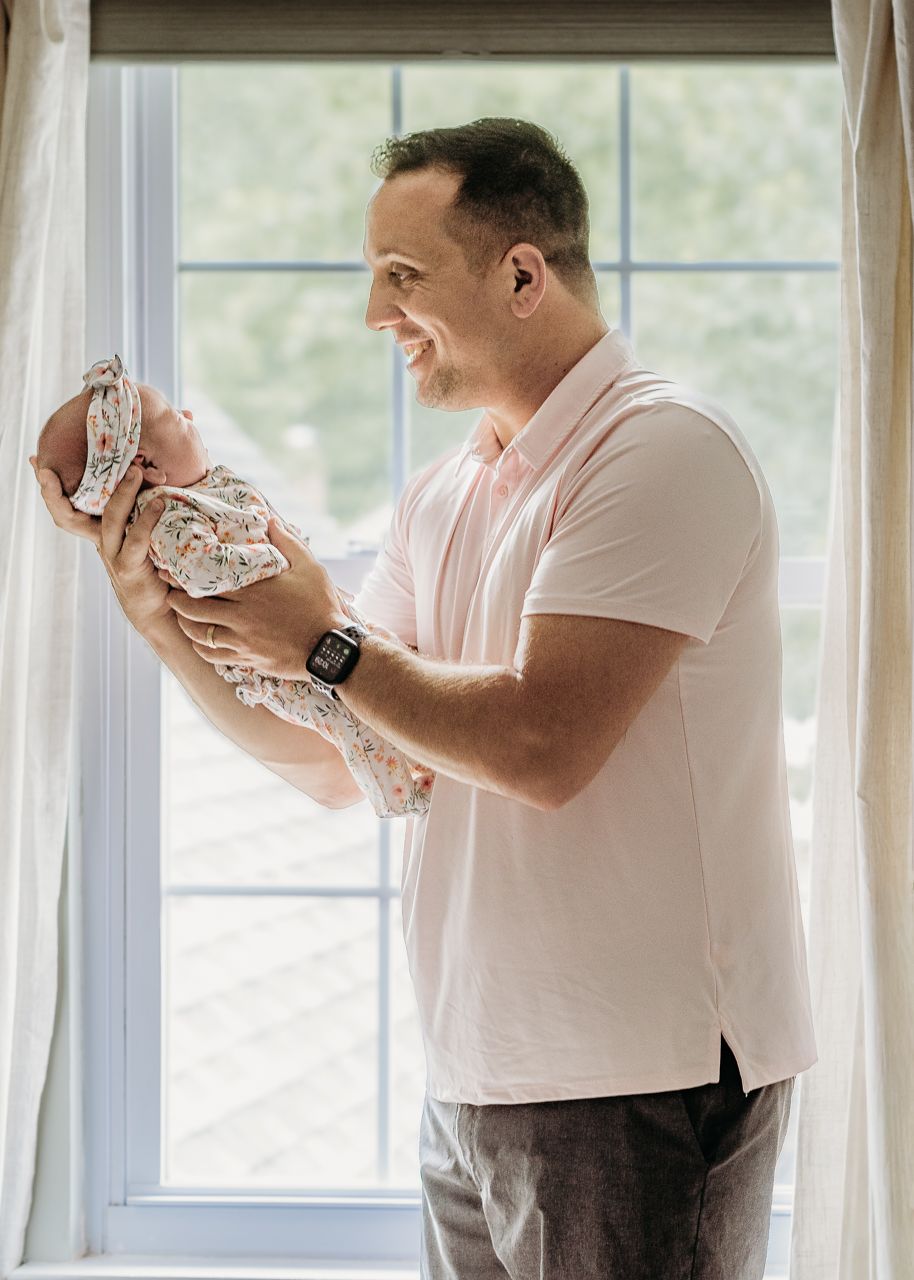 Dad smiling while holding his newborn daughter by a bright window.