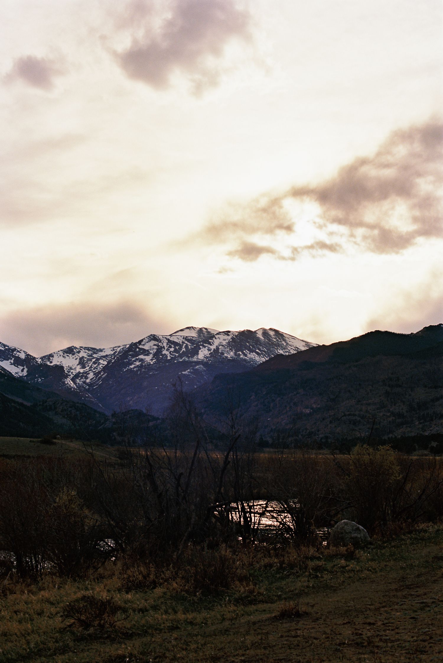 Snow-capped mountain range at sunset with dramatic clouds, green hills, and a serene valley landscape.