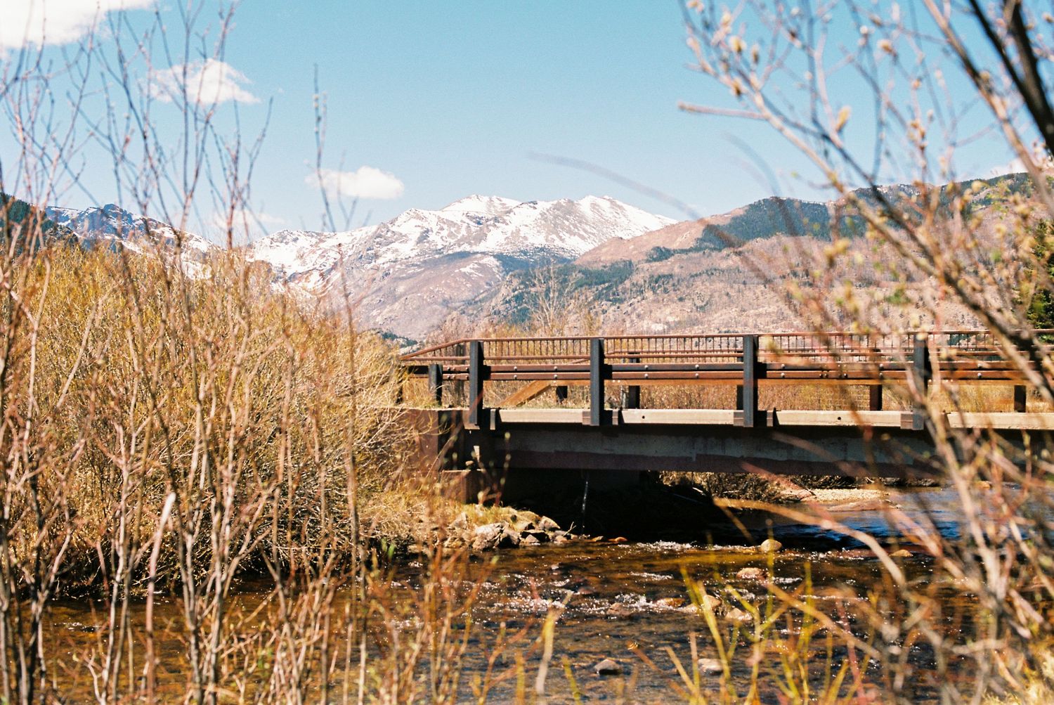 Wooden bridge over a stream with snow-capped mountains and autumn shrubs in a scenic landscape.