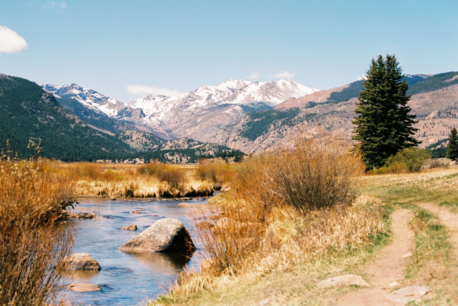 A scenic mountain stream flows past golden shrubs and a dirt path with snow-capped peaks in the background.