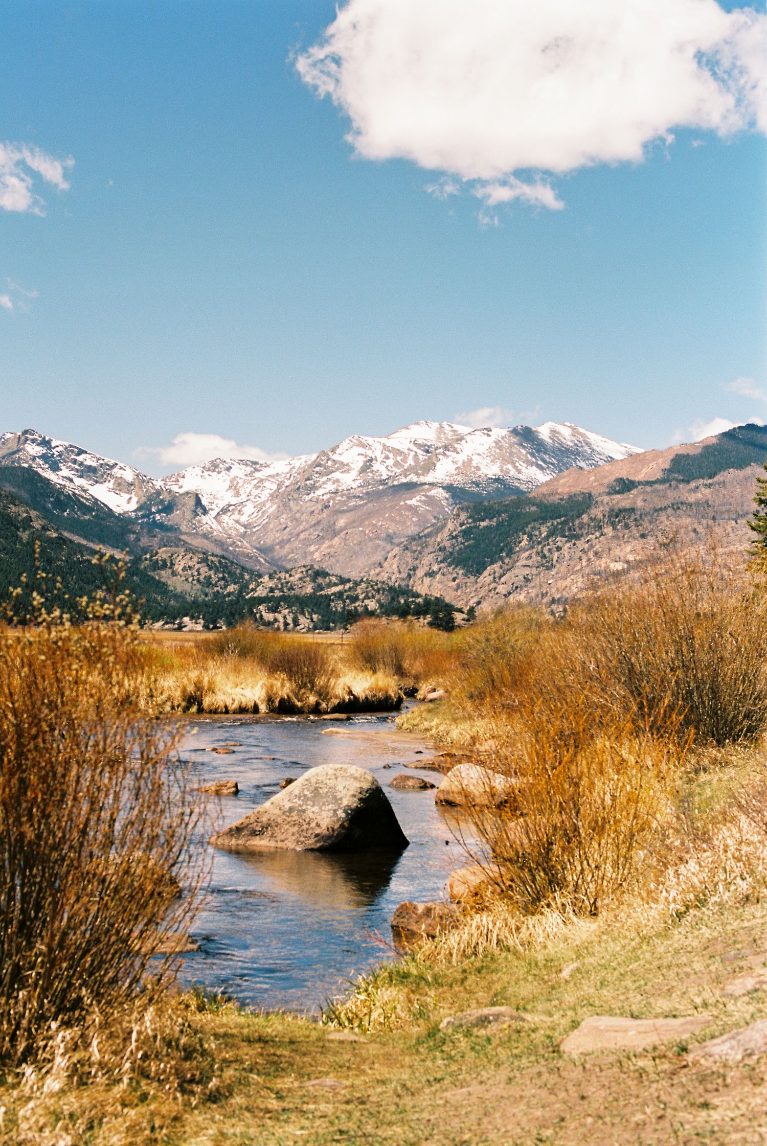 A rocky mountain stream flows through golden autumn brush with snow-capped peaks under a blue sky.