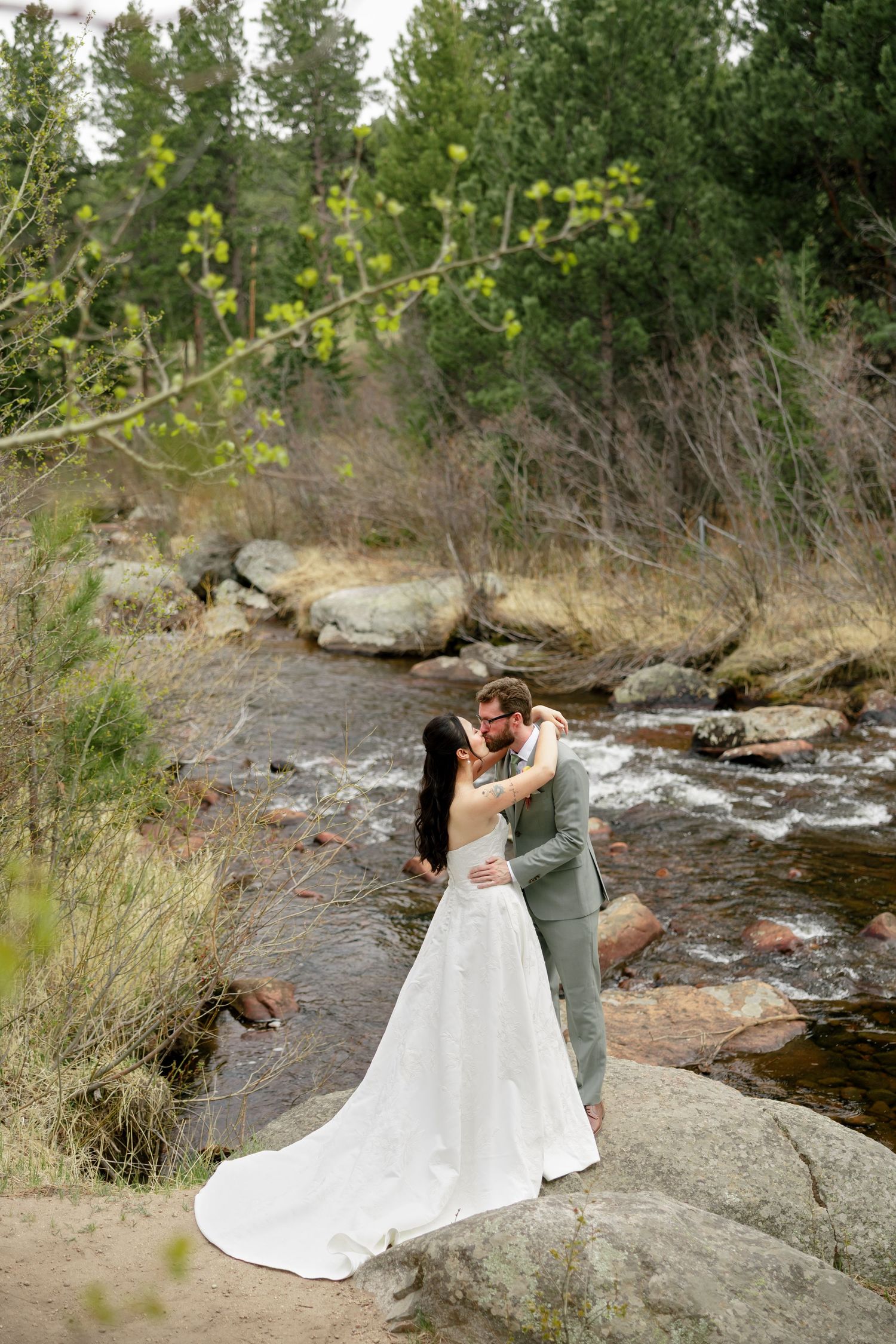 Bride and groom embrace romantically by a rocky mountain stream surrounded by lush green forest on their wedding day.