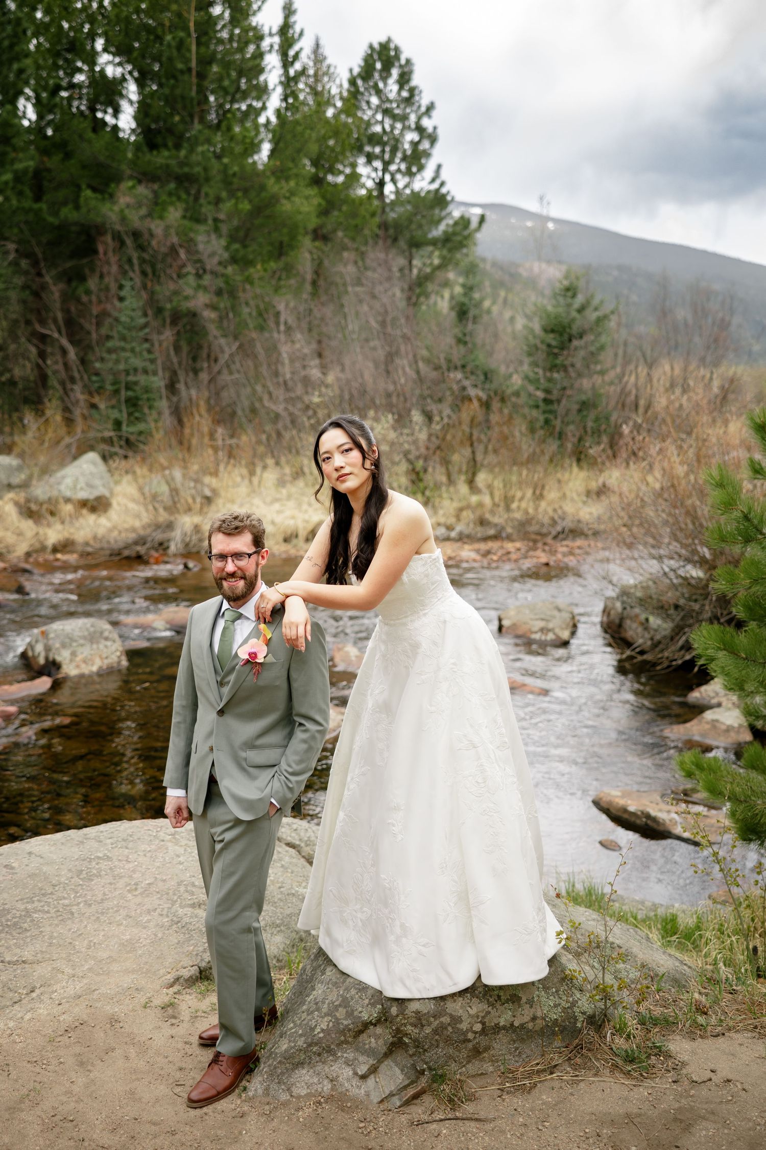 Bride in white gown stands on rock beside groom in gray suit near mountain stream surrounded by pine trees.