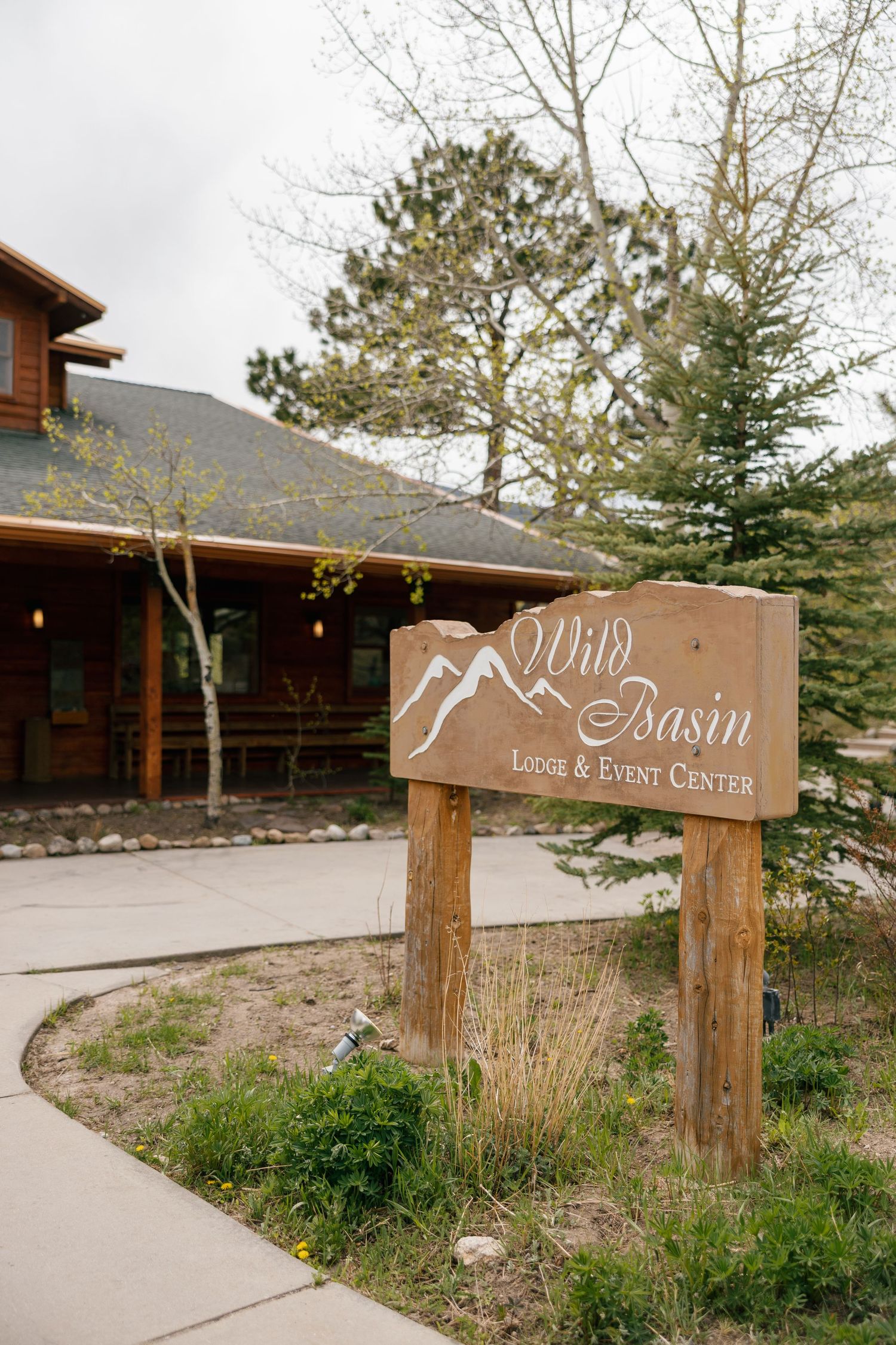 Wooden sign for Wild Basin Lodge and Event Center surrounded by pine trees and rustic log building in Colorado.