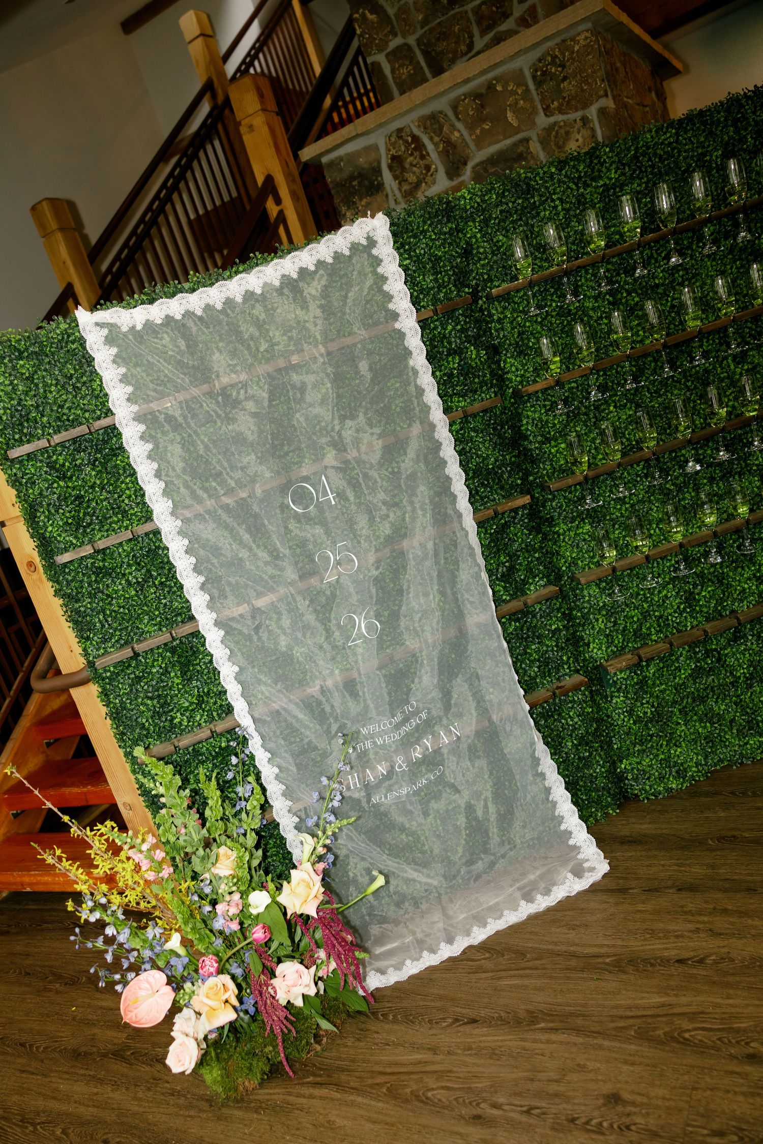 Elegant wedding seating chart on sheer fabric with lace trim, displayed against a green boxwood wall with floral arrangement.