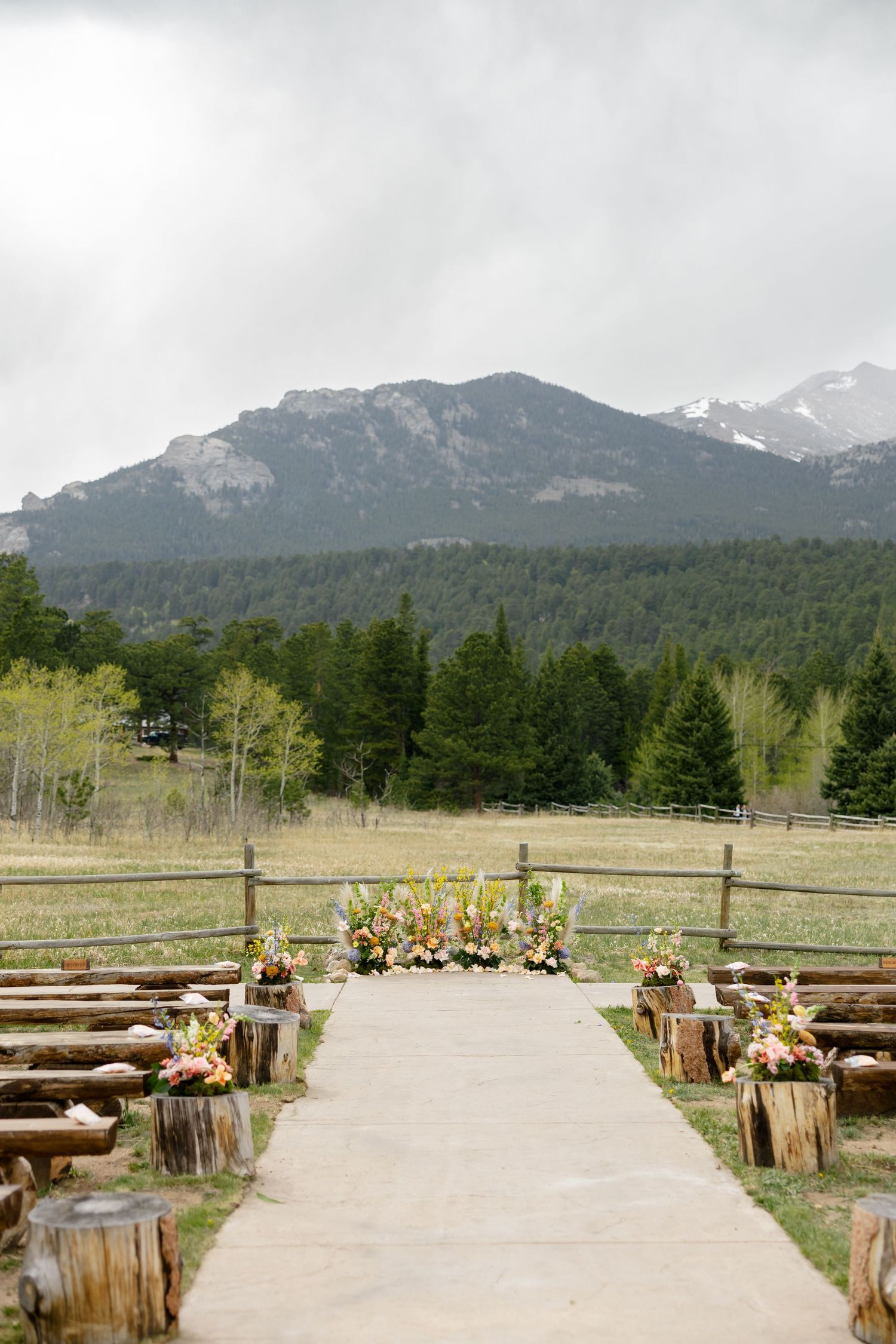 Outdoor mountain wedding ceremony aisle with rustic log seating, floral arrangements, and a stunning Rocky Mountain backdrop.