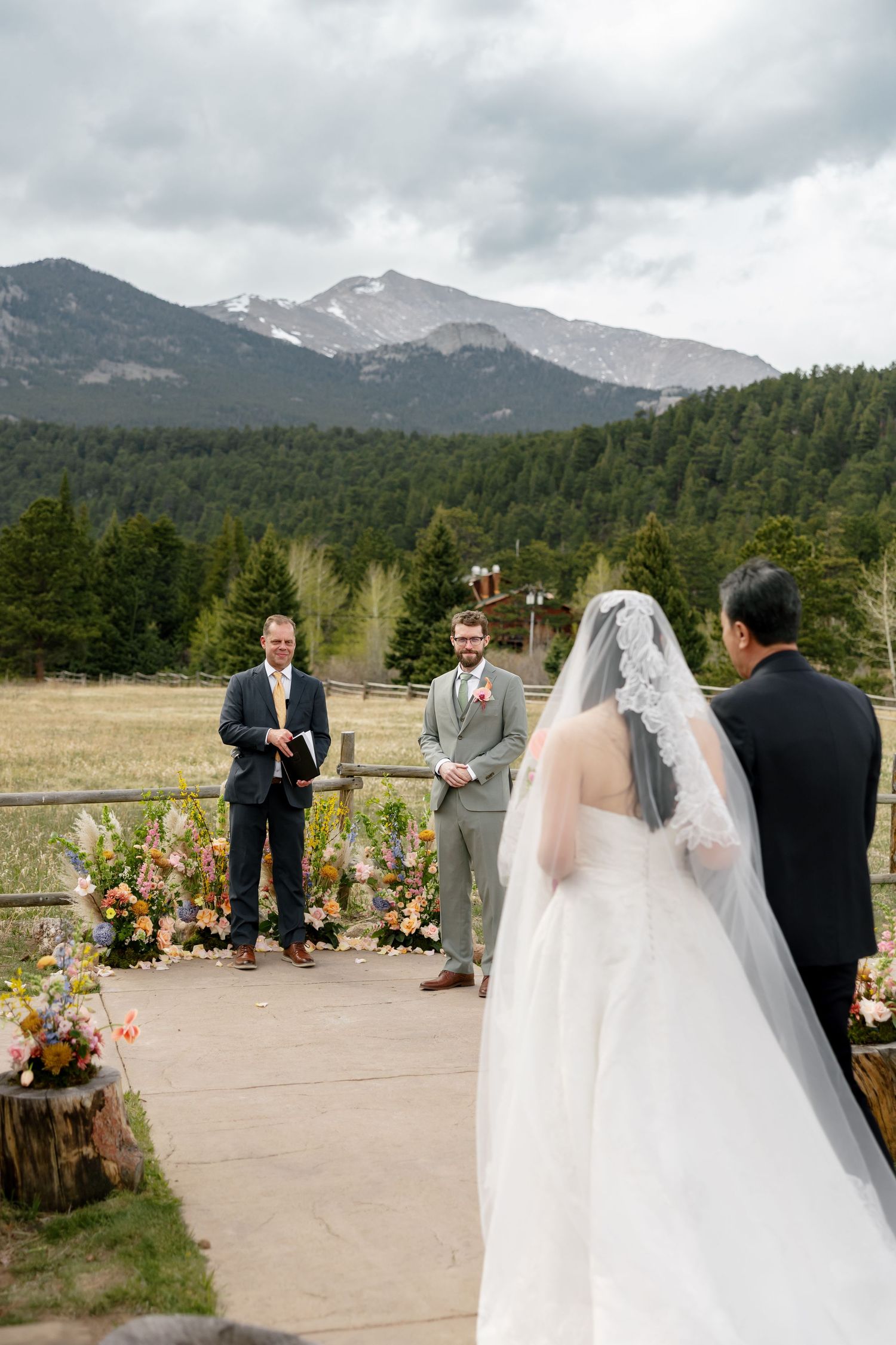 Outdoor mountain wedding ceremony with bride in lace veil, groom in gray suit, and officiant at floral altar in Colorado.