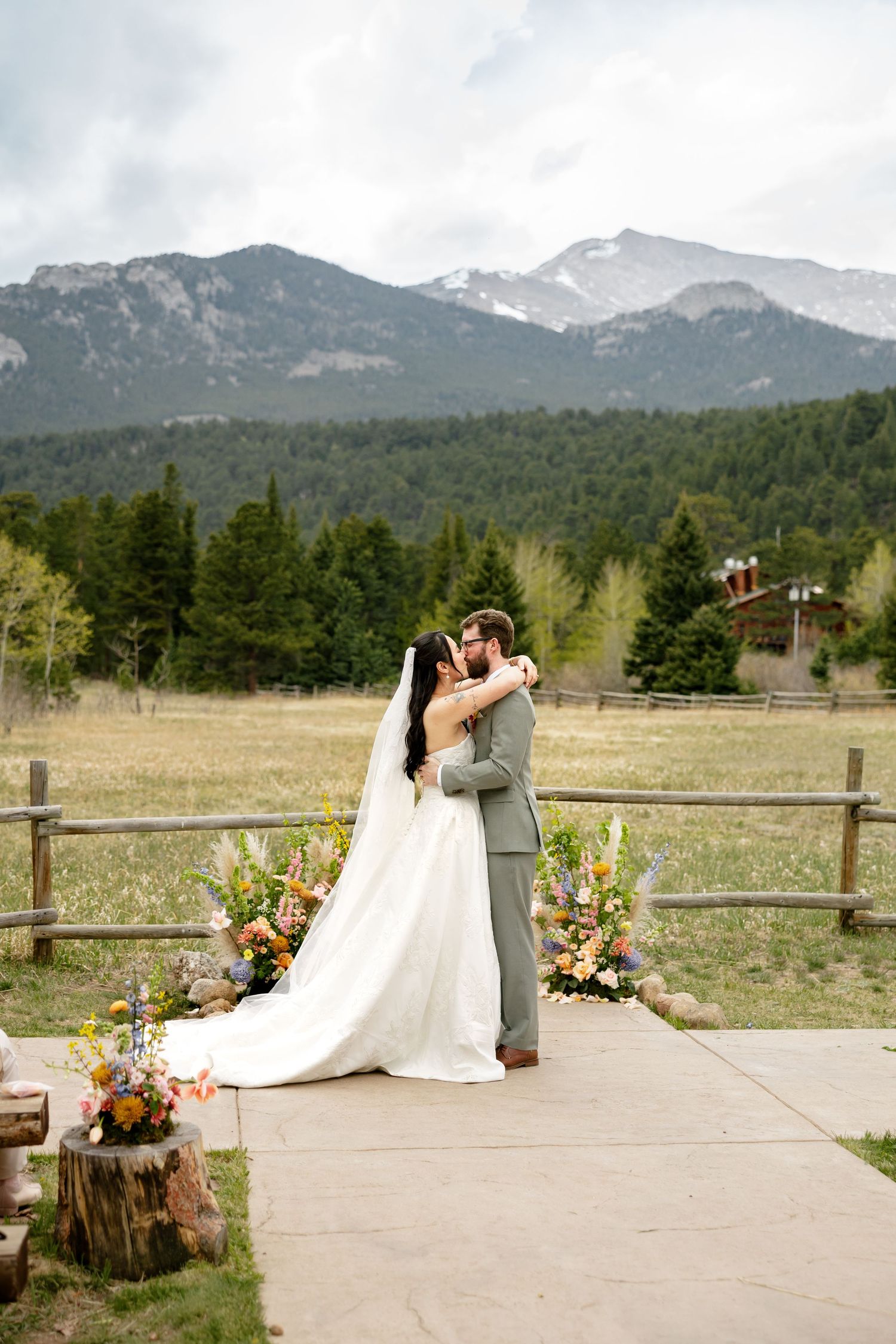 Bride and groom share first kiss at outdoor mountain wedding ceremony with colorful floral arrangements and snow-capped peaks backdrop.