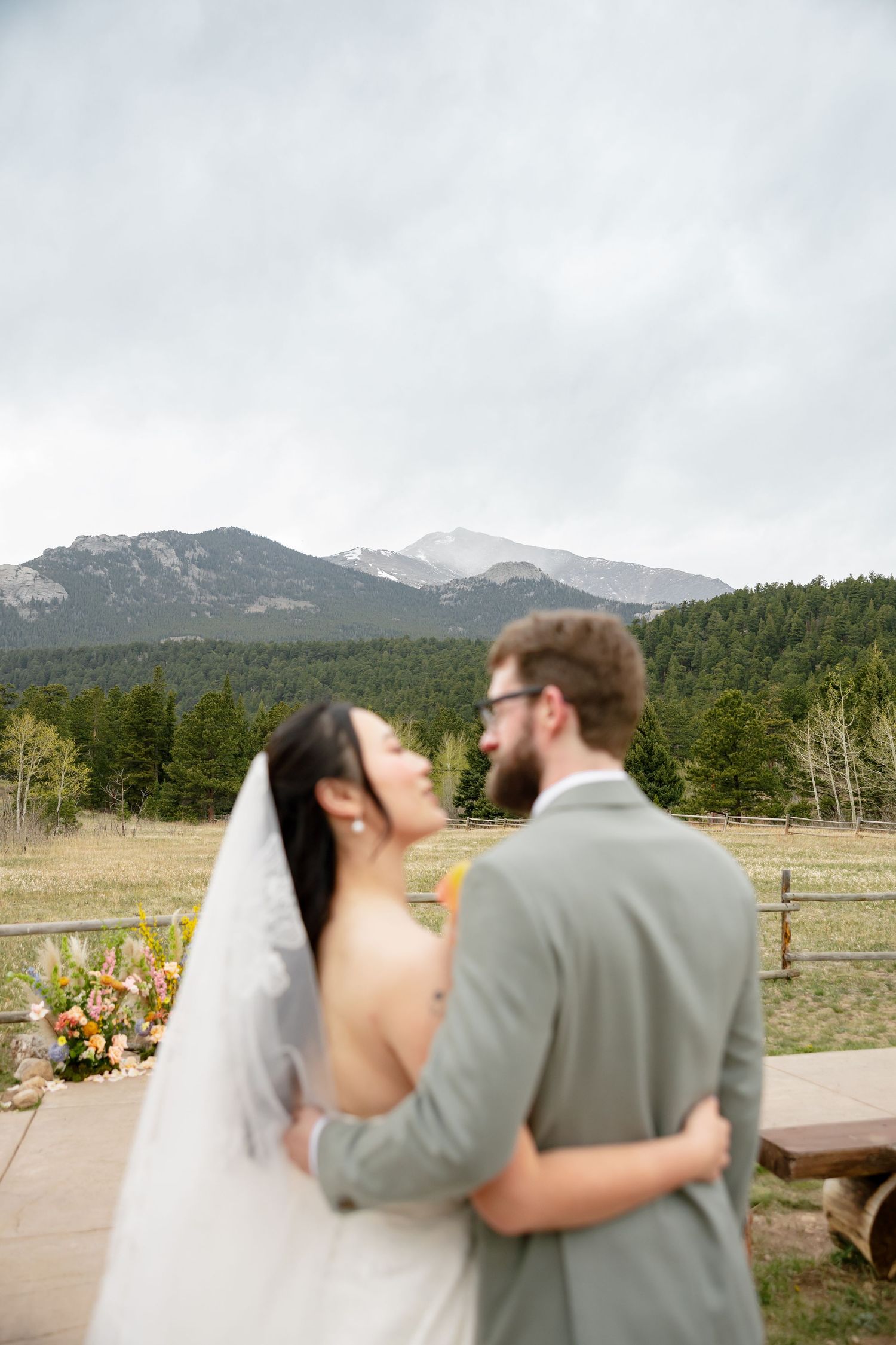 A bride and groom embrace outdoors with a stunning mountain and forest backdrop on their wedding day.