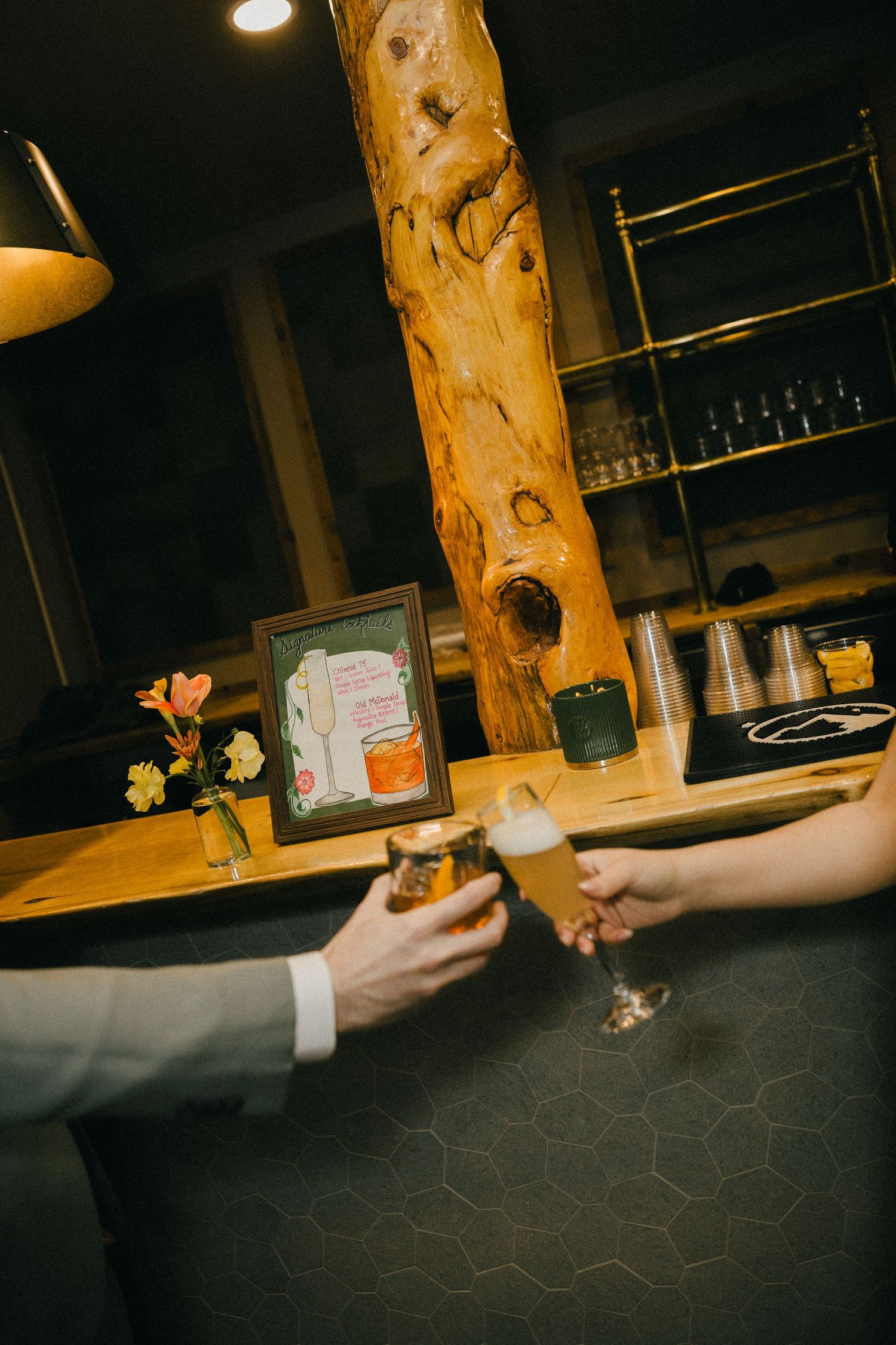 Couple toasting champagne glasses at rustic wooden bar with natural log pillar during wedding reception.