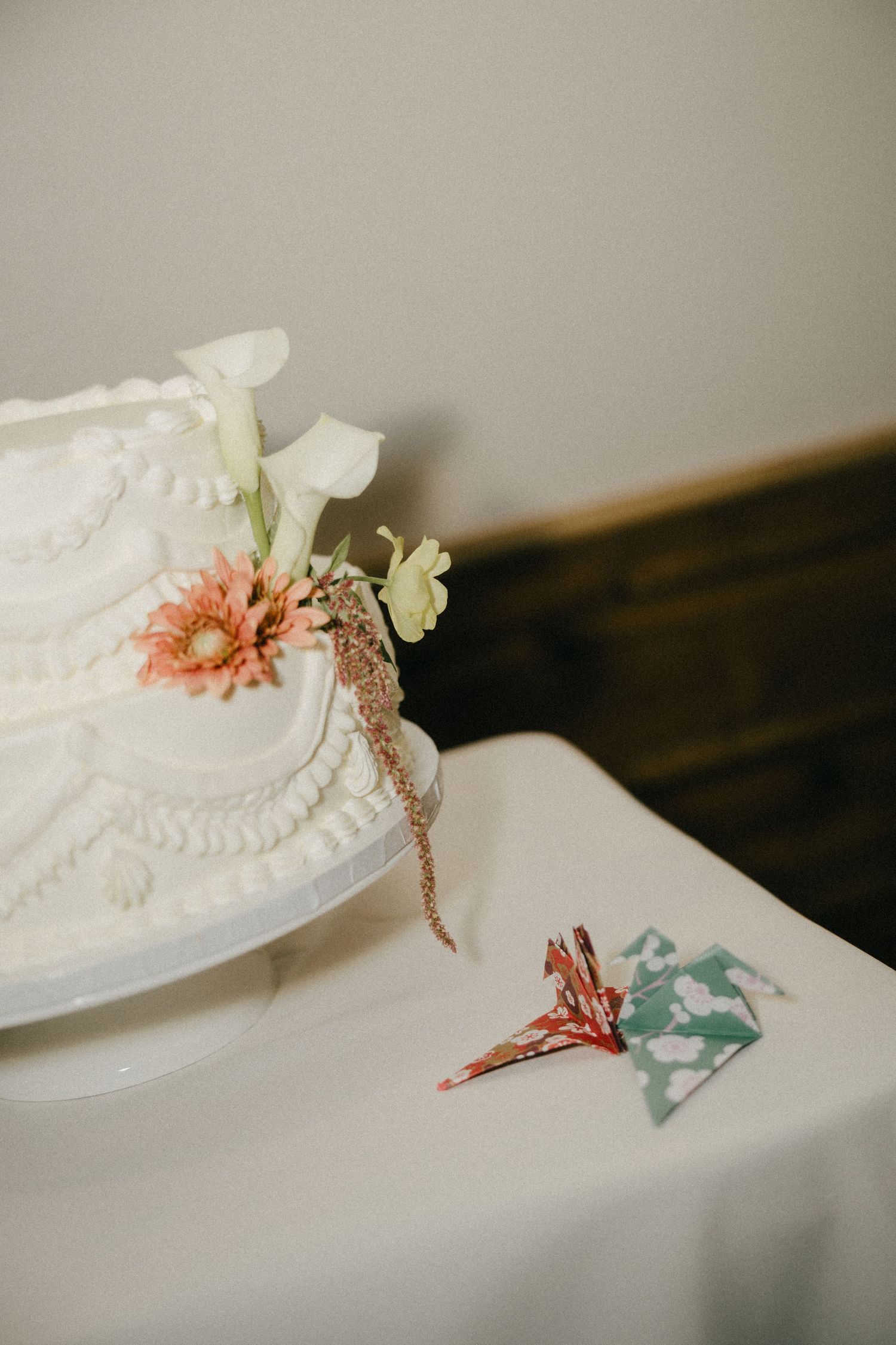 White tiered wedding cake adorned with pink gerbera daisy and flowers, beside colorful origami crane on white tablecloth.