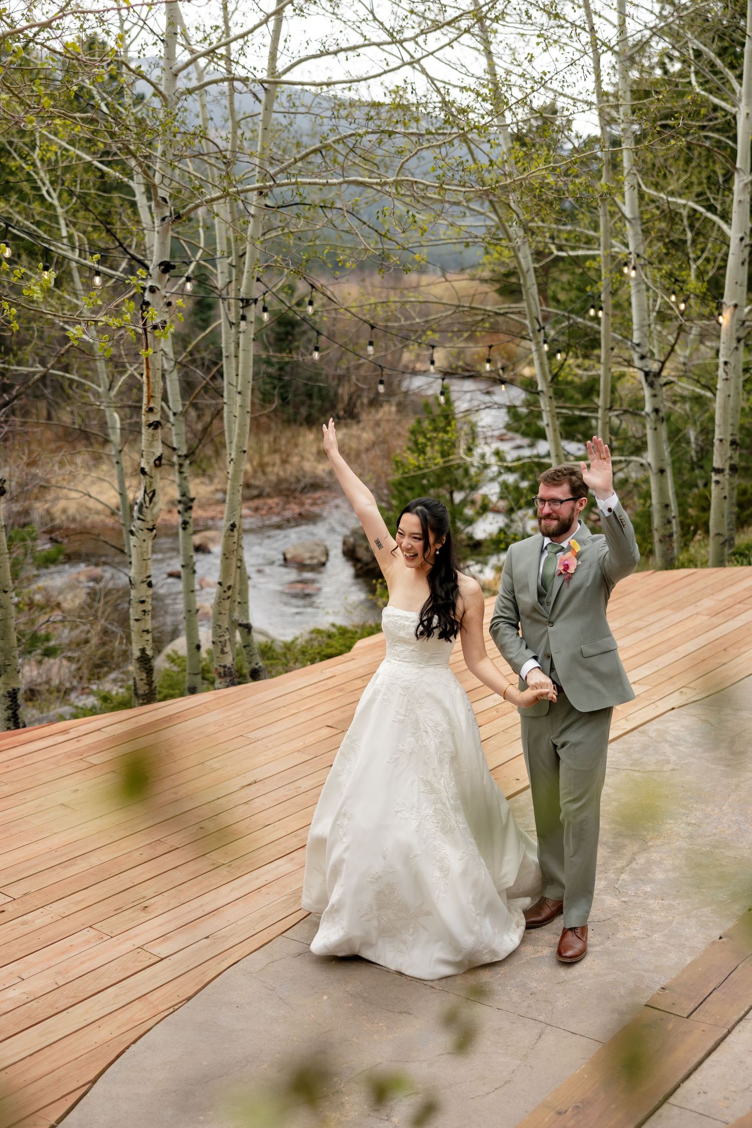 Joyful bride and groom celebrating on wooden deck surrounded by aspen trees and mountain river backdrop.